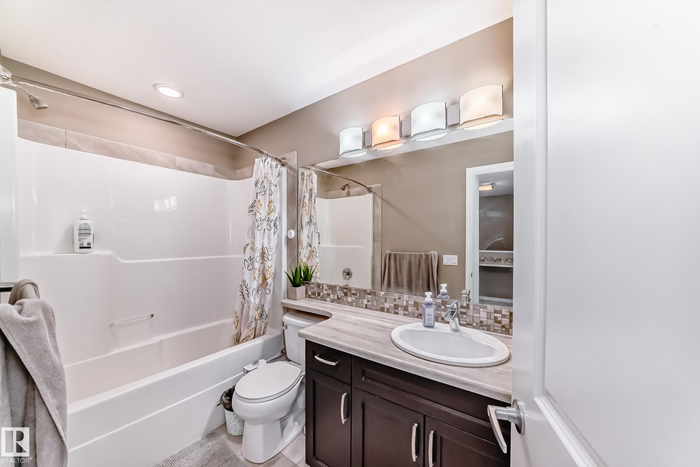 Bathroom featuring a white tub and shower combination, dark wood-finish vanity with a light-toned countertop, mosaic tile backsplash, and a large mirror with a four-light fixture - 5020 Orchards Gate, Edmonton, AB - Indoor Photo Showing Bathroom