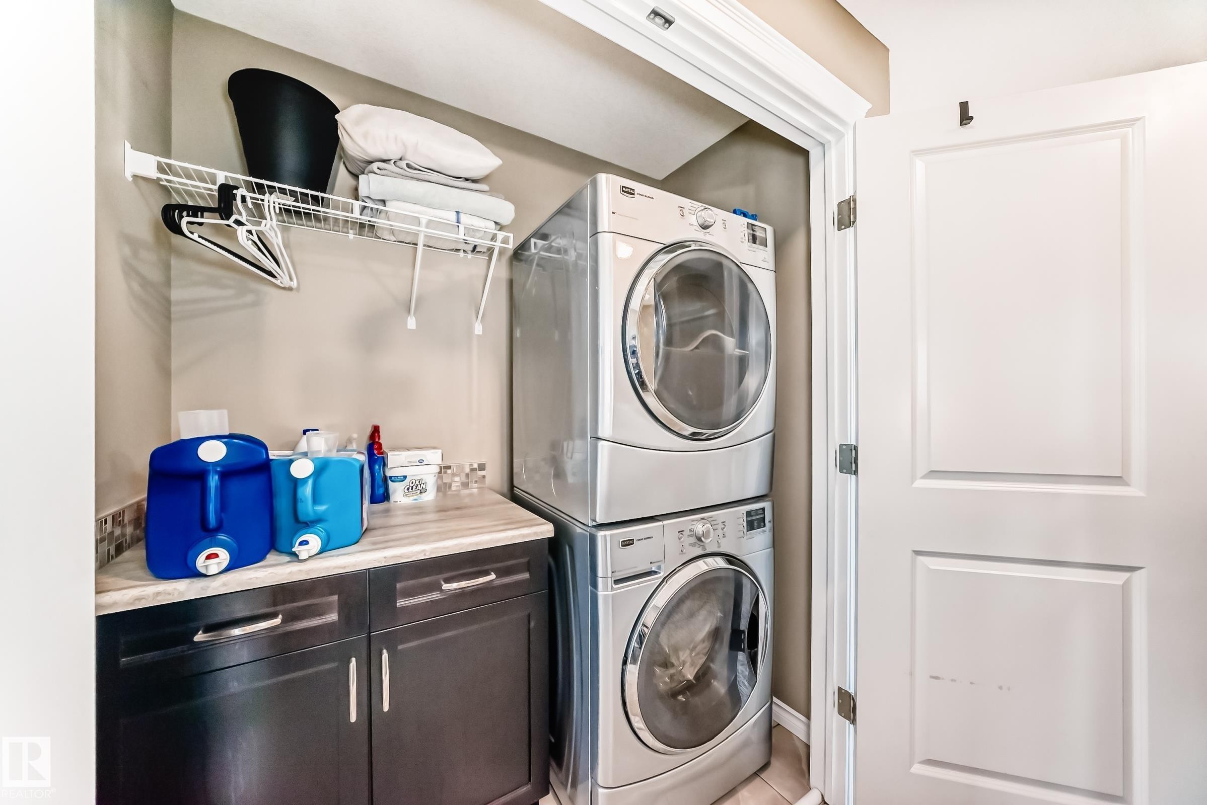 Laundry area featuring a stacked washer and dryer, dark wood-finish cabinetry with silver hardware, a light-toned countertop, and a wire shelving unit - 5020 Orchards Gate, Edmonton, AB - Indoor Photo Showing Laundry Room
