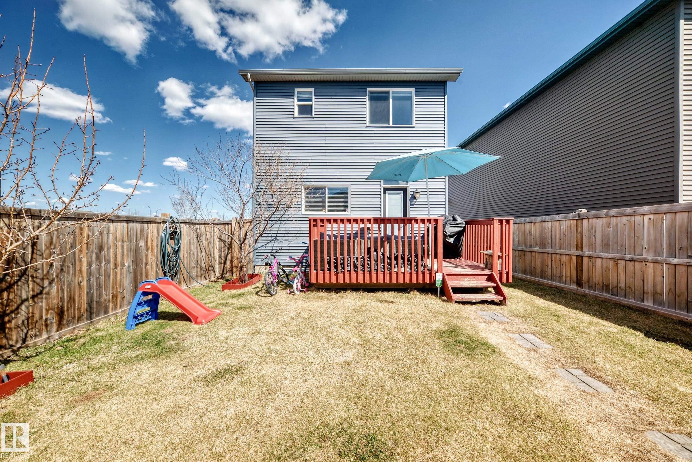 Rear exterior featuring light blue horizontal siding, a red wood-finish deck with steps, a full wood-finish privacy fence, and a grass lawn with a stone paver path - 5020 Orchards Gate, Edmonton, AB - Outdoor With Deck Patio Veranda