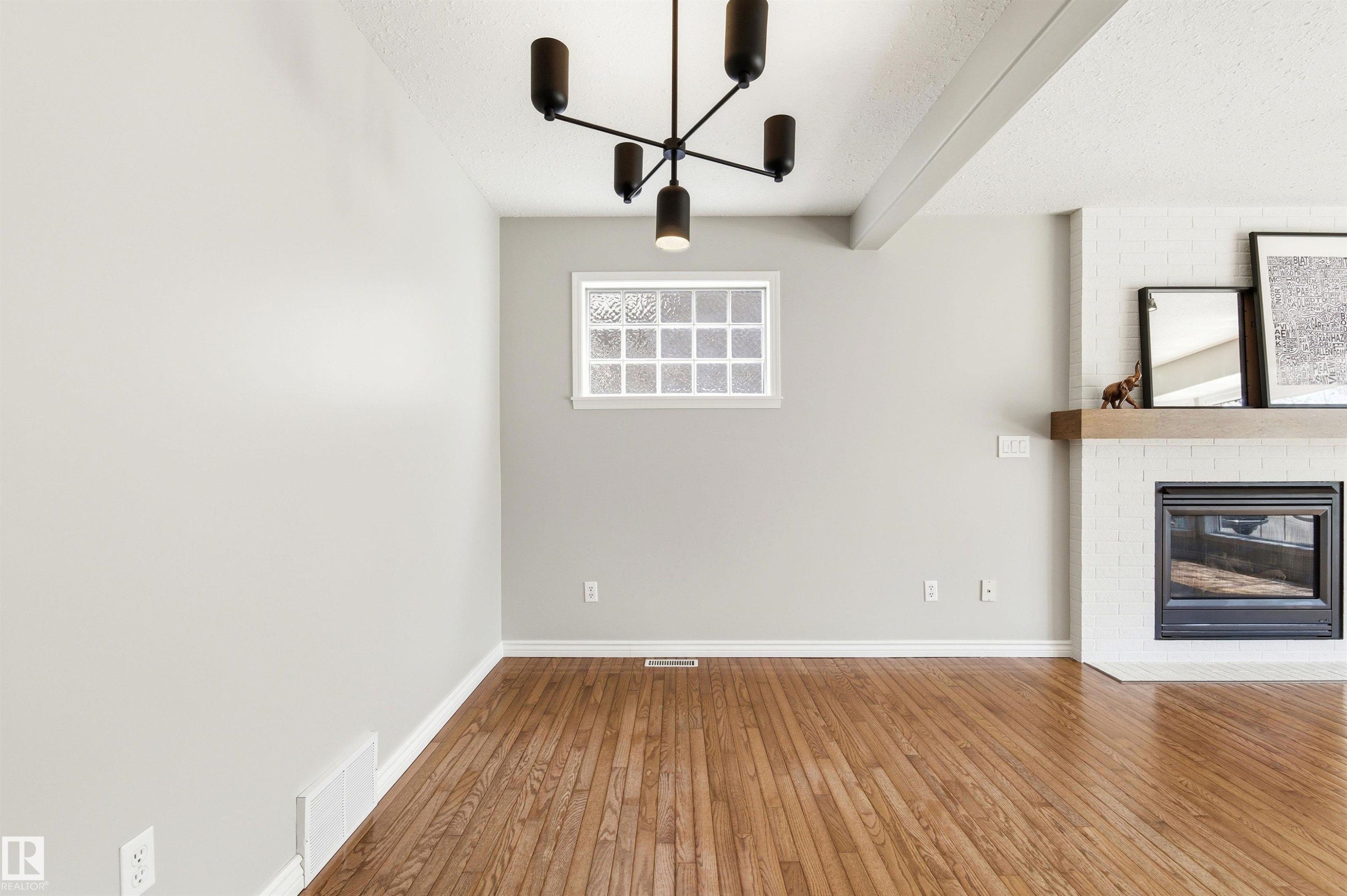 Spacious room featuring wood-finish flooring and a white brick fireplace with a wood mantel - 8730 92A Avenue, Edmonton, AB - Indoor Photo Showing Other Room With Fireplace
