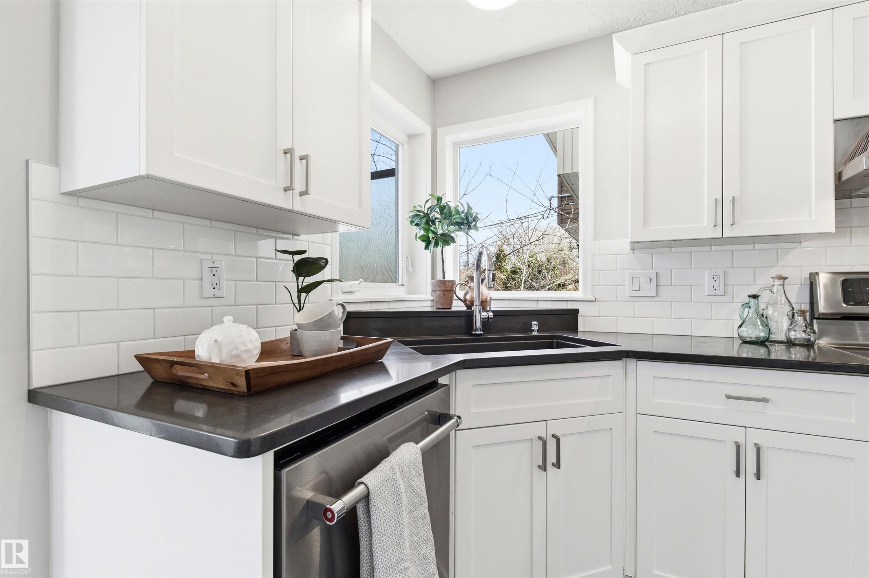 Corner kitchen sink with a stainless steel faucet, dark countertop, white subway tile backsplash, white shaker-style cabinetry, and stainless steel dishwasher - 8730 92A Avenue, Edmonton, AB - Indoor Photo Showing Kitchen With Double Sink
