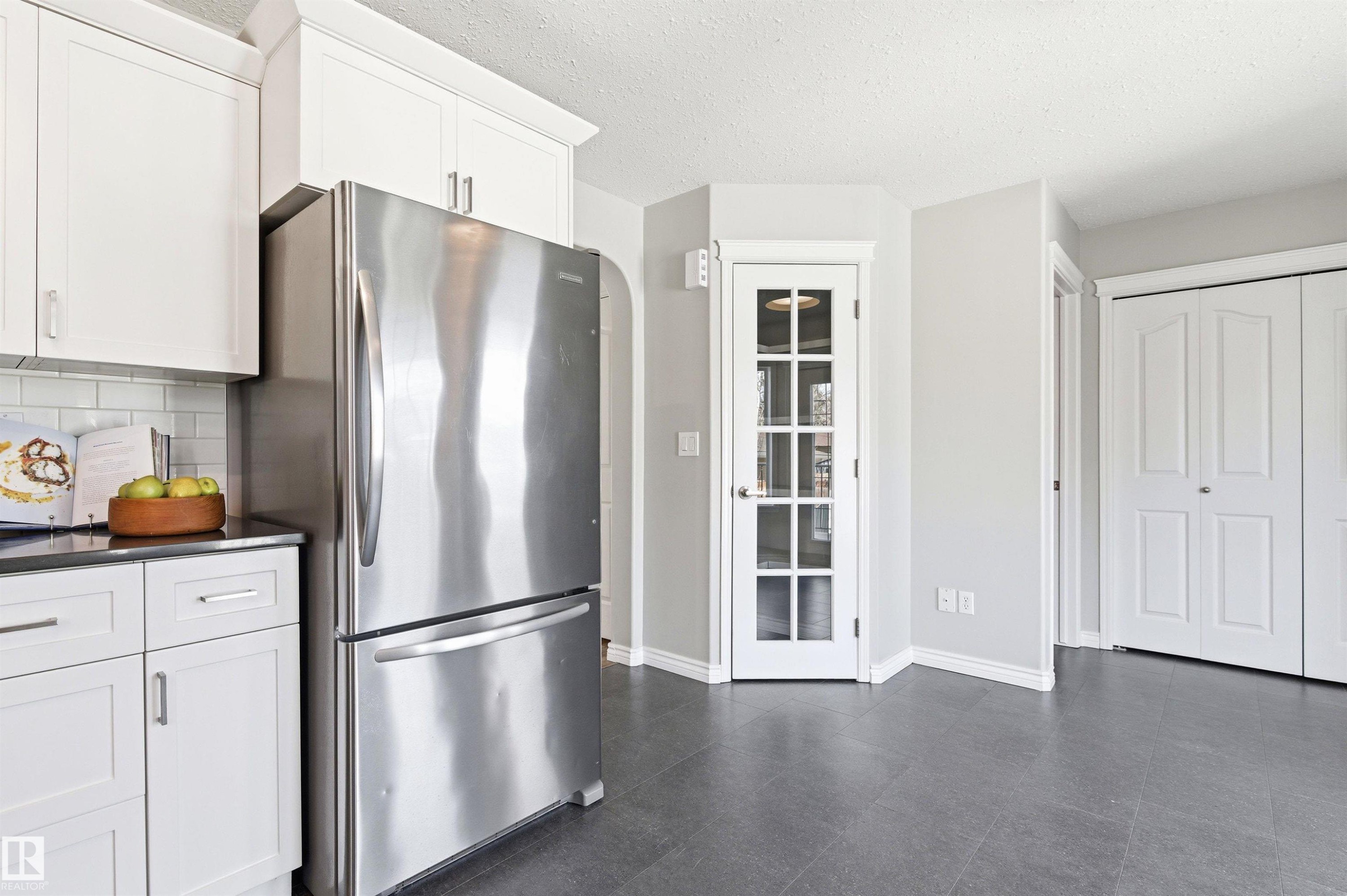 Kitchen area featuring white shaker-style cabinetry with chrome hardware, a stainless steel refrigerator, and dark countertops - 8730 92A Avenue, Edmonton, AB - Indoor Photo Showing Kitchen