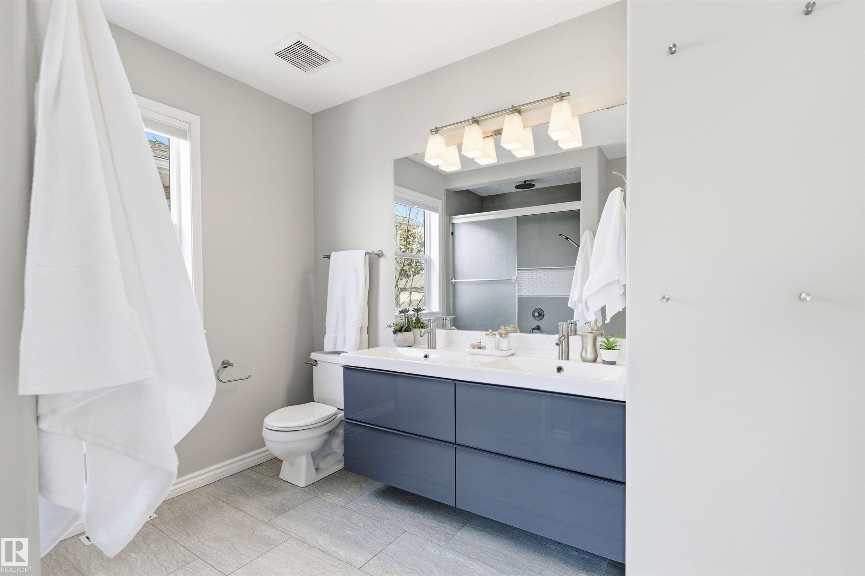 Bathroom featuring a double vanity with a white countertop and blue cabinetry, a contemporary four-light fixture, a toilet, a window, and large format tile flooring - 8730 92A Avenue, Edmonton, AB - Indoor Photo Showing Bathroom