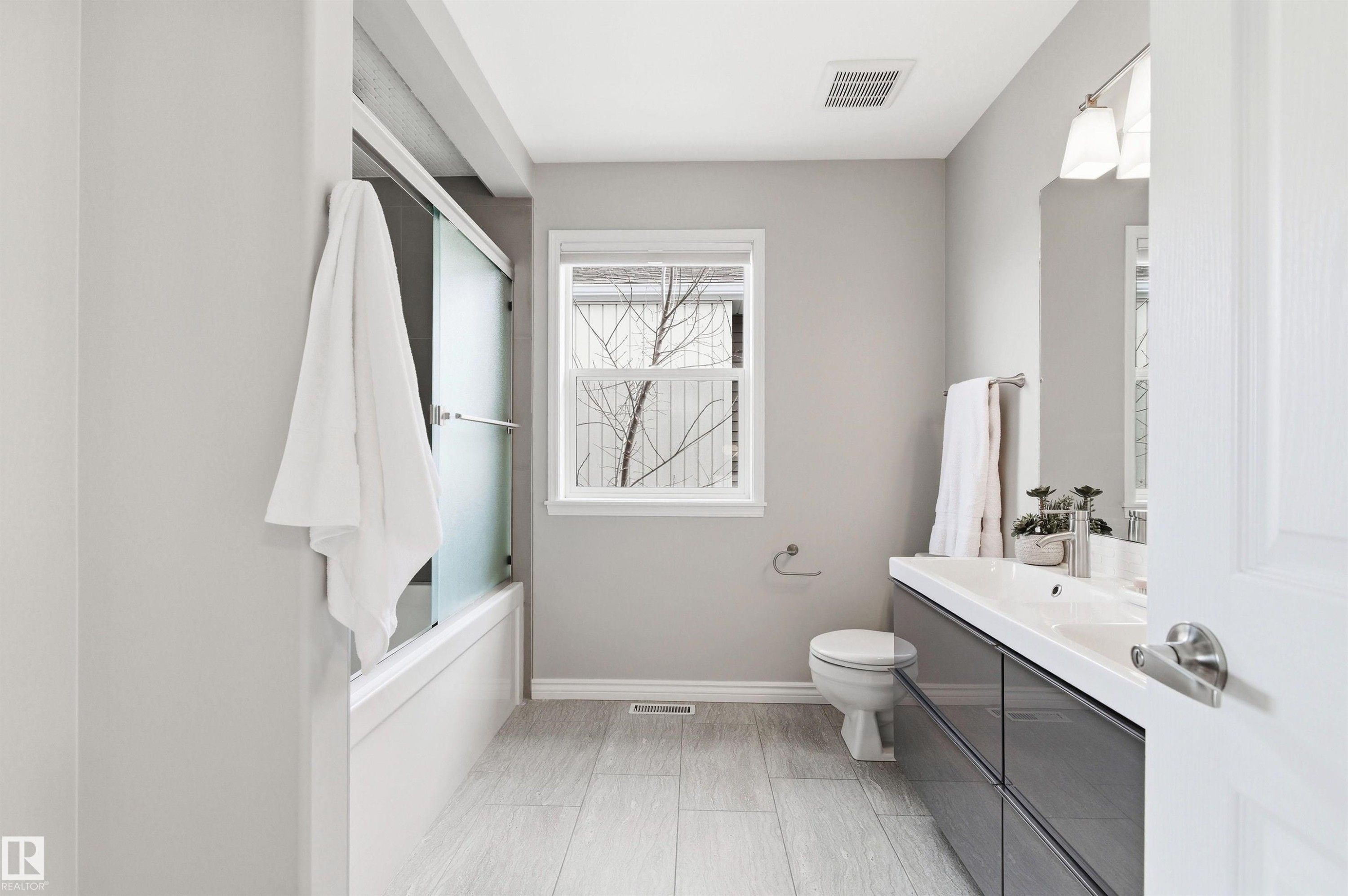 Bathroom featuring a vanity with a white countertop and dark gray cabinetry, a toilet, a window with white trim, a shower-tub combination with a sliding frosted glass door, and light wood-finish flooring - 8730 92A Avenue, Edmonton, AB - Indoor Photo Showing Bathroom