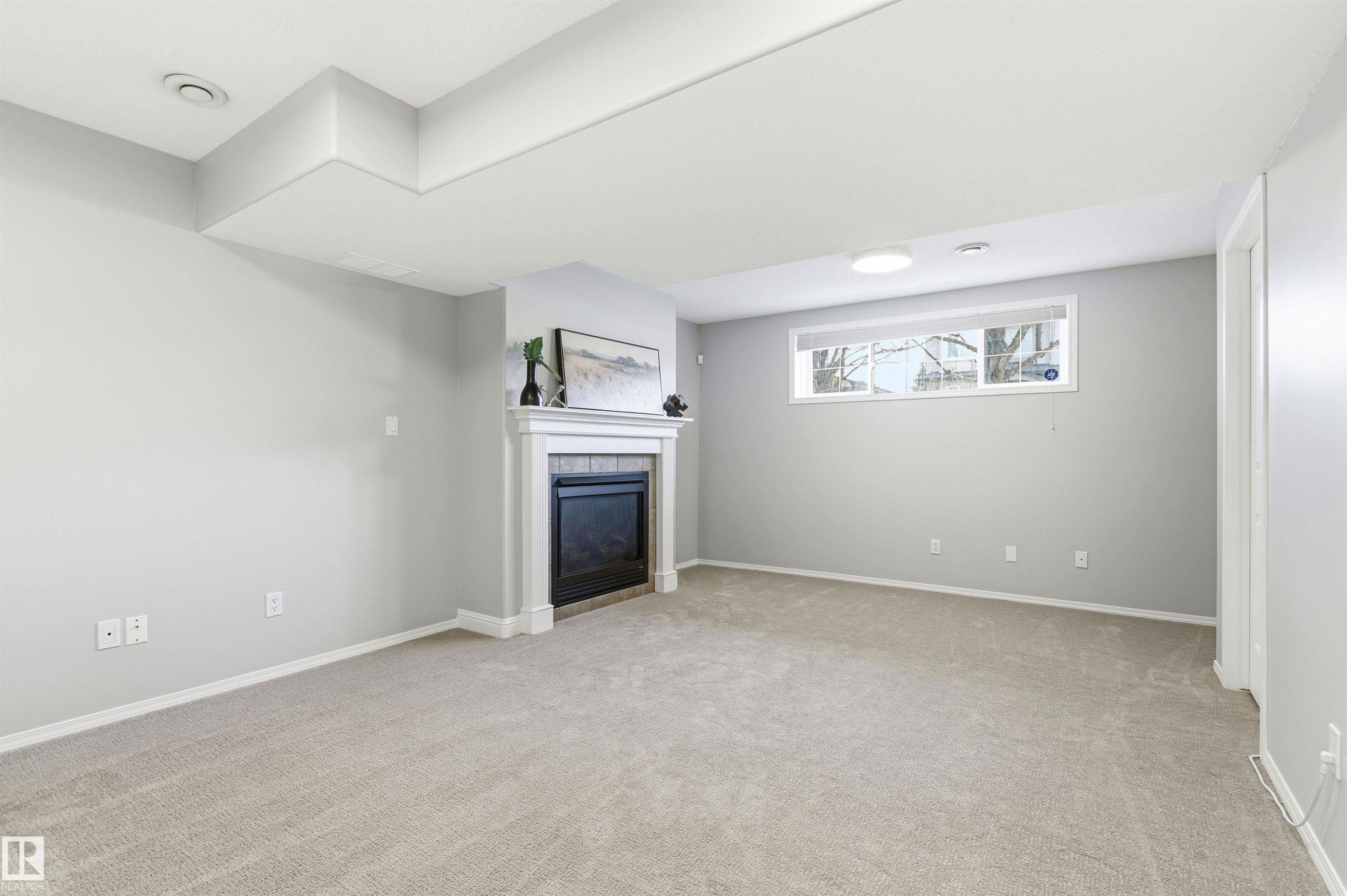 Spacious room featuring a white-mantled fireplace, neutral gray walls, light-toned carpeting, and a window providing natural light - 8730 92A Avenue, Edmonton, AB - Indoor Photo Showing Other Room