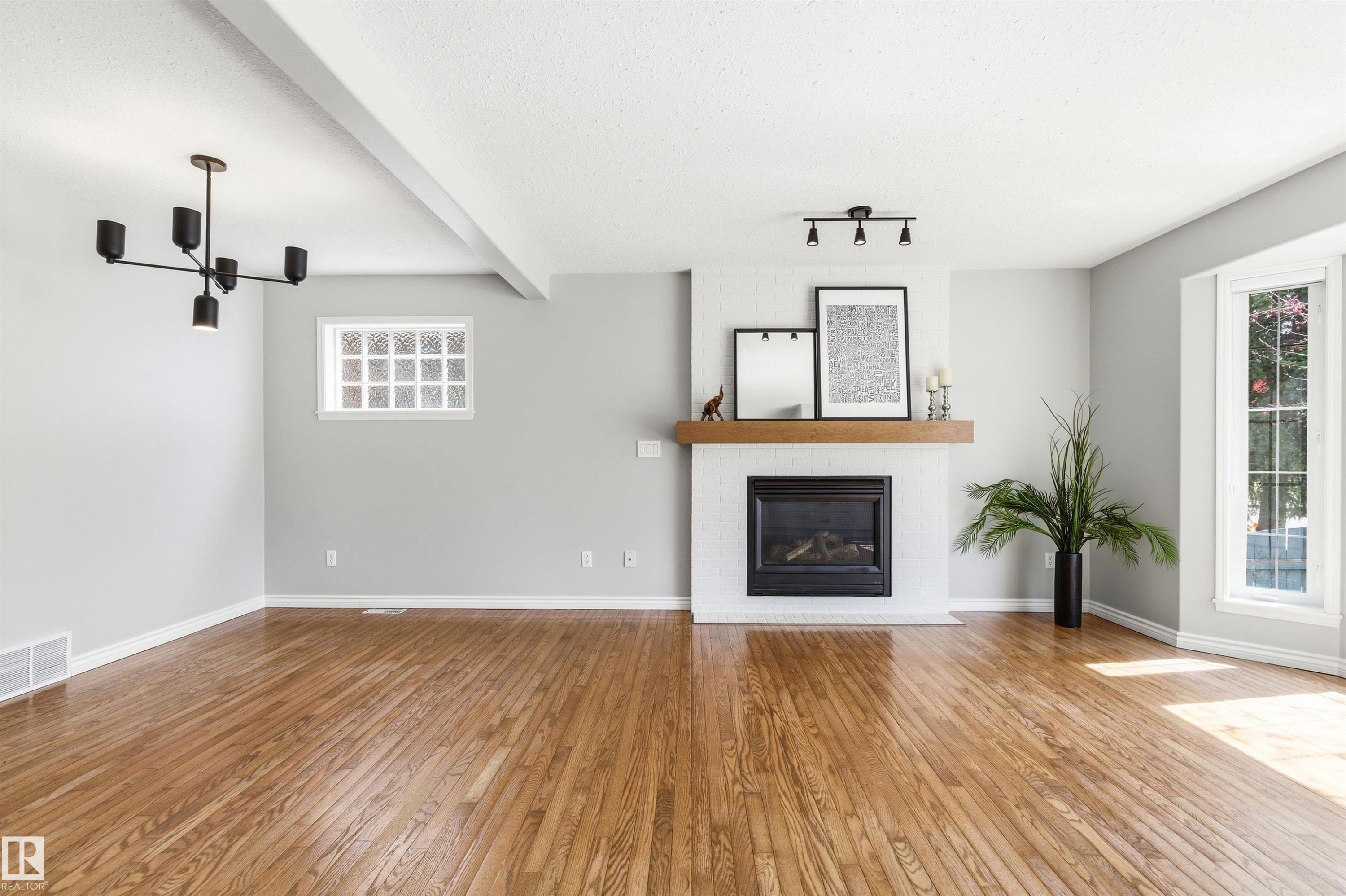 Expansive room featuring wood-finish flooring, a white brick-style fireplace with a wood mantel, and a black built-in fireplace insert - 8730 92A Avenue, Edmonton, AB - Indoor Photo Showing Living Room With Fireplace