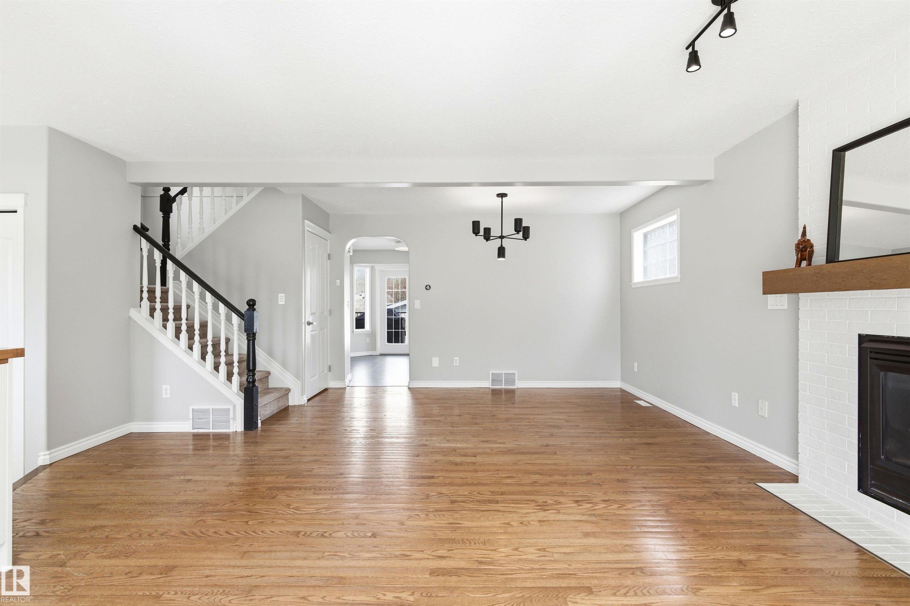 Open living area featuring wood-finish flooring, a contemporary black chandelier, and a white brick fireplace with a wood mantle - 8730 92A Avenue, Edmonton, AB - Indoor With Fireplace