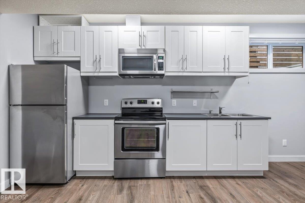 Kitchen featuring stainless steel appliances, white shaker-style cabinetry, dark countertops, a double basin sink, and wood-finish flooring - 8706 88 Avenue, Edmonton, AB - Indoor Photo Showing Kitchen With Stainless Steel Kitchen With Double Sink