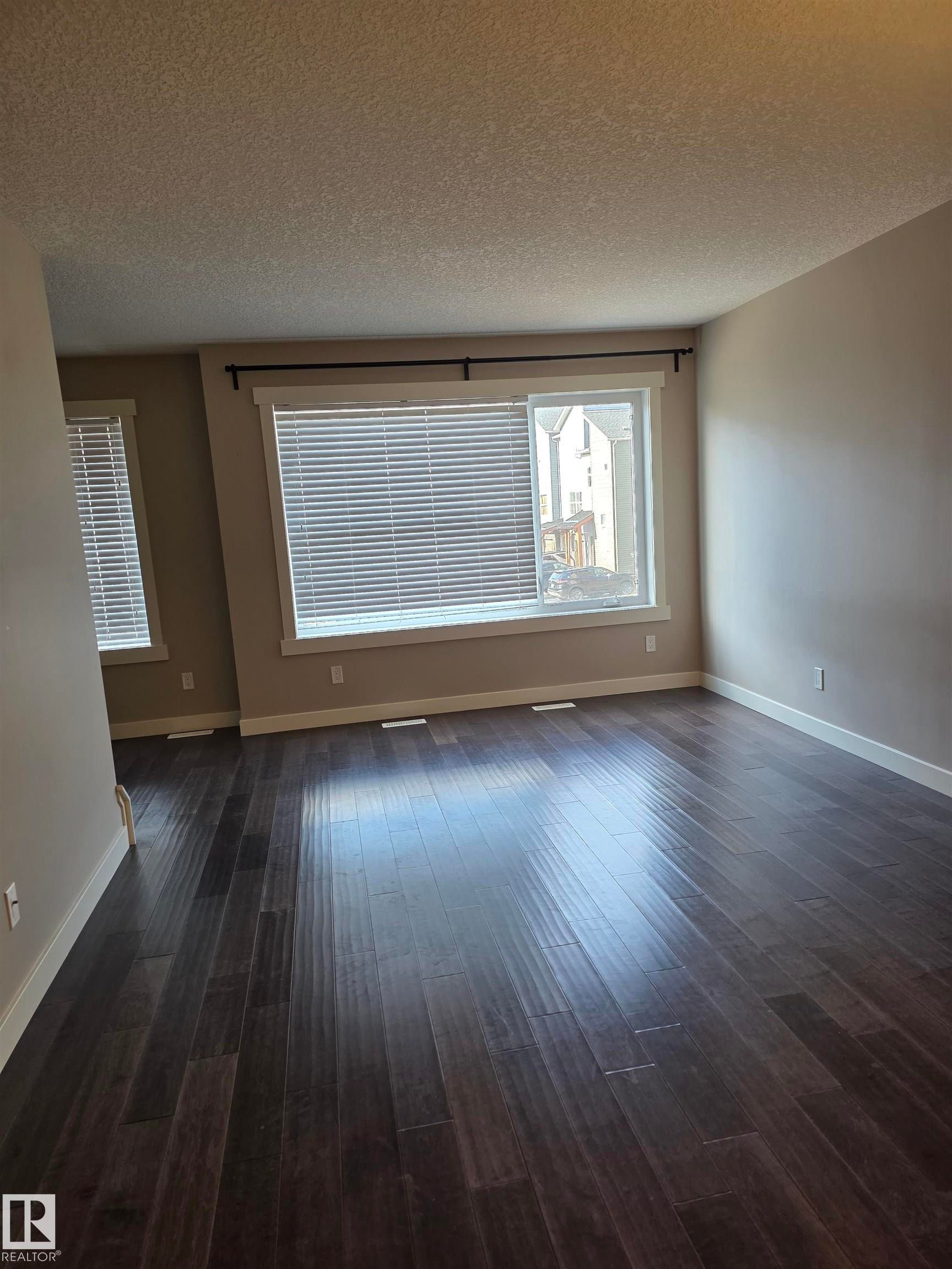 Spacious room featuring dark wood-finish flooring, two large windows with blinds, and neutral painted walls - 295 401 Southfork, Leduc, AB - Indoor Photo Showing Other Room