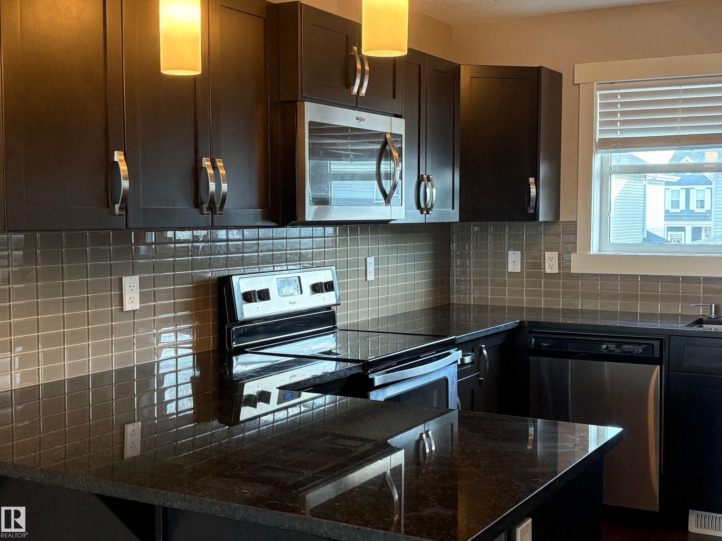 Kitchen featuring dark cabinetry, dark countertops, square tile backsplash, stainless steel appliances, and pendant lighting - 295 401 Southfork, Leduc, AB - Indoor Photo Showing Kitchen With Upgraded Kitchen