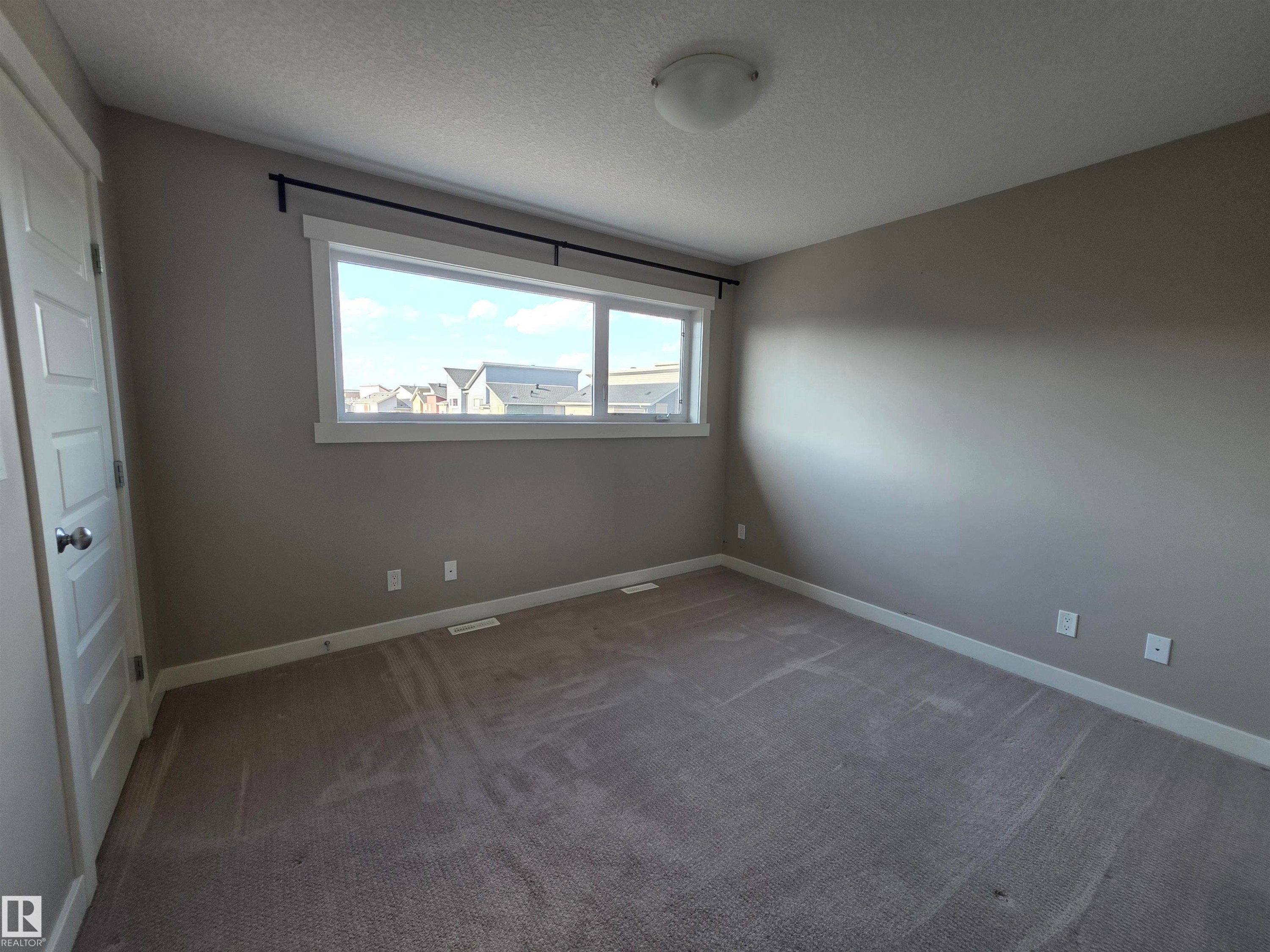 Neutral-toned room featuring a wide window with white trim, gray wall paint, and gray carpet flooring - 295 401 Southfork, Leduc, AB - Indoor Photo Showing Other Room