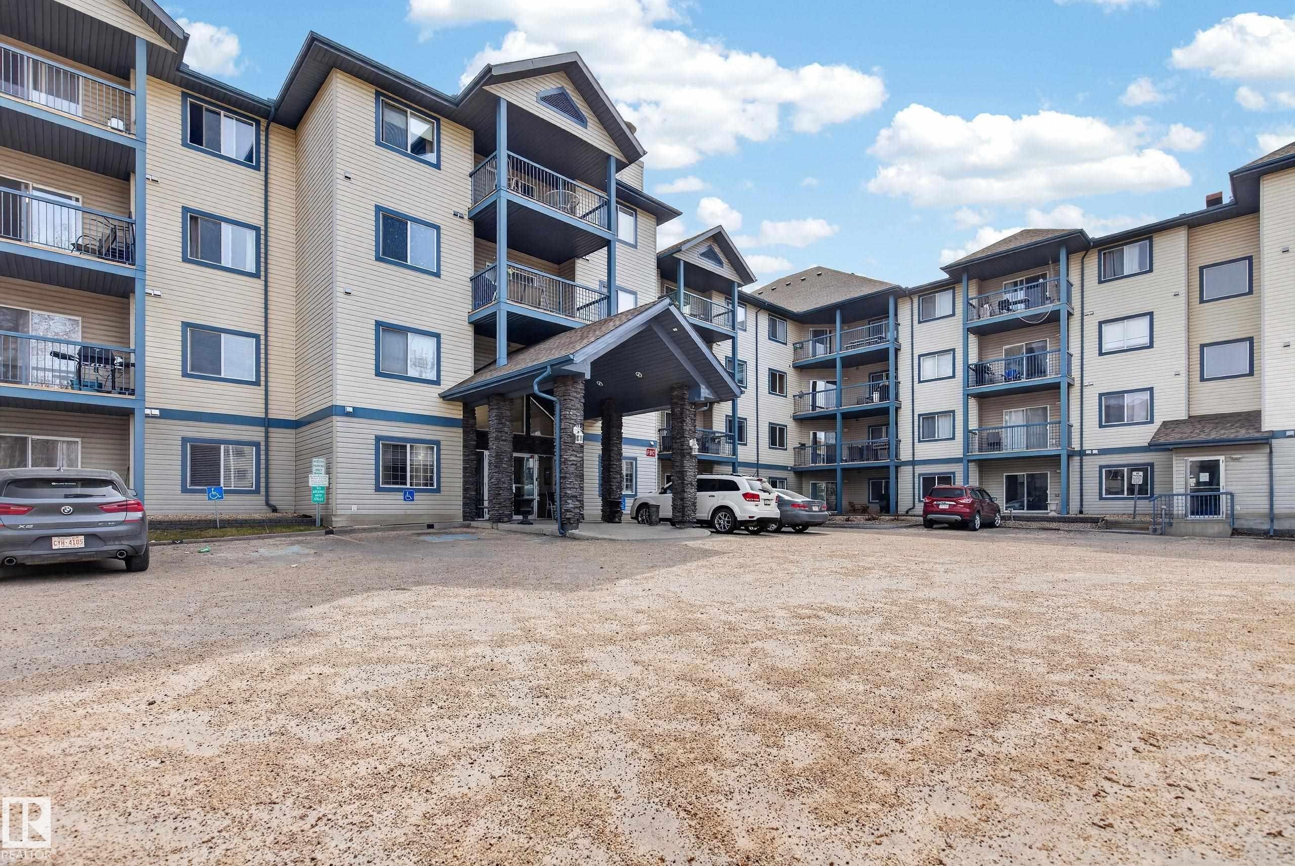 Multi-story residential building featuring light-colored siding with dark trim, multiple balconies with glass railings, and a prominent covered entrance with stone accents - #319 16303 95 St Nw, Edmonton, AB - Outdoor With Facade