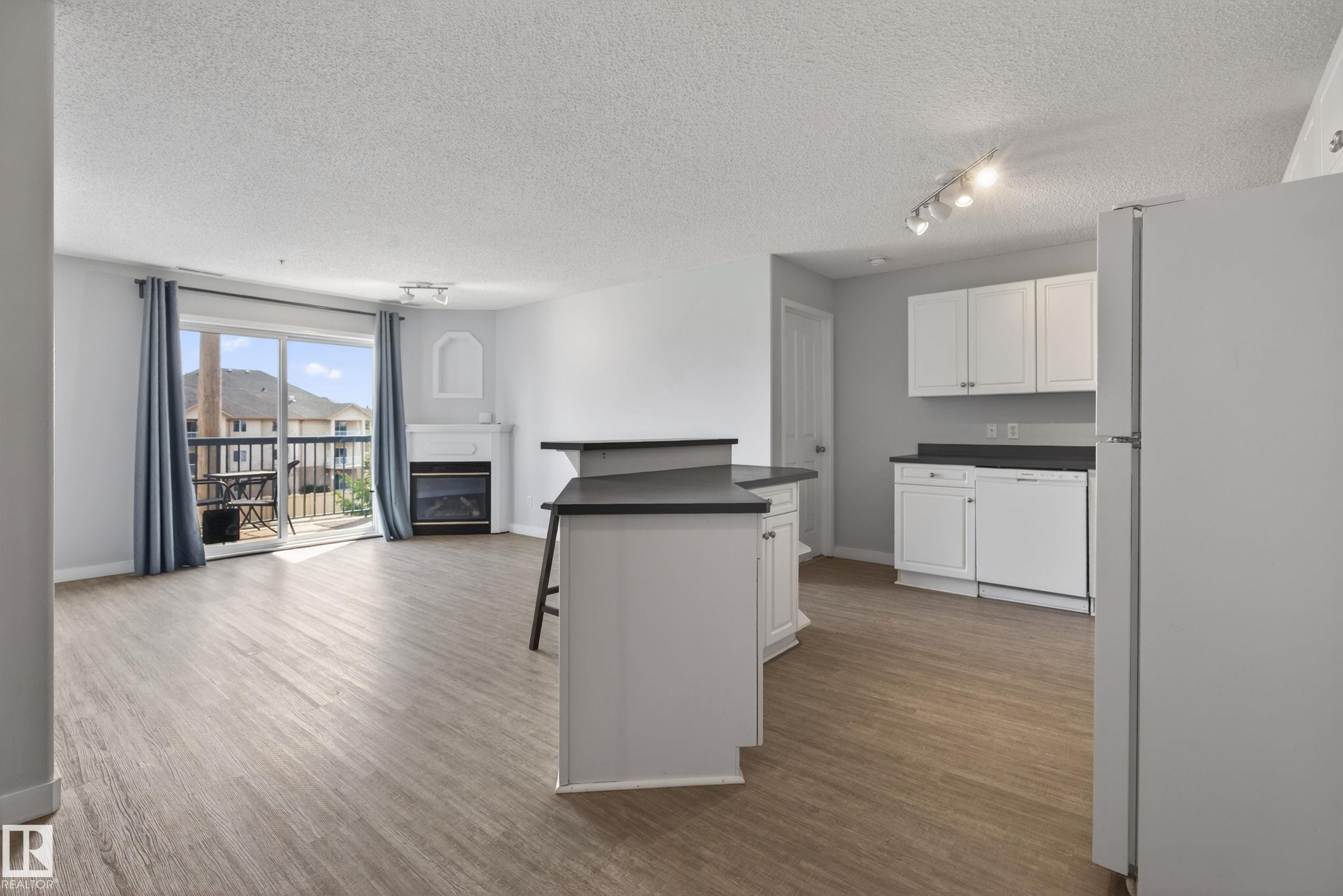 Open-concept living space featuring wood-finish flooring, a fireplace with a mantel, a balcony with a black railing, and a kitchen with white cabinetry and dark countertops - #319 16303 95 St Nw, Edmonton, AB - Indoor