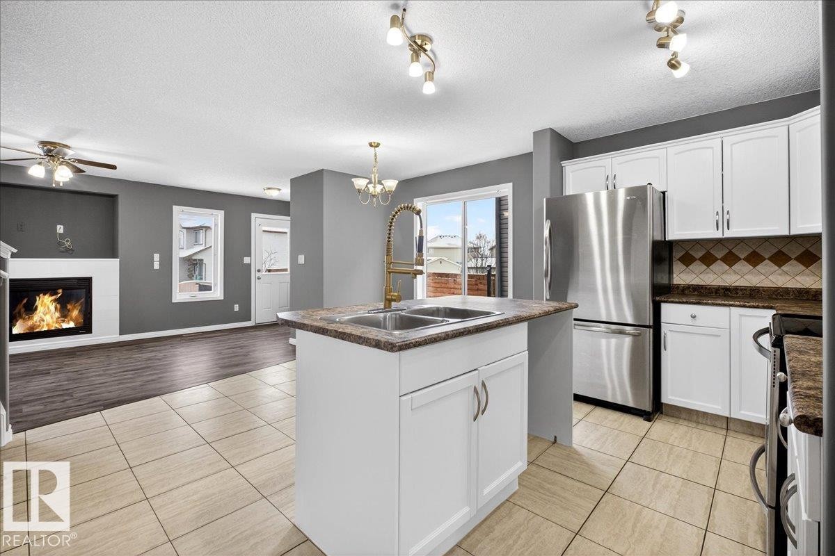 Kitchen featuring white cabinetry, stainless steel appliances, a glass covered fireplace, a textured ceiling, and open floor plan - 150 Acacia Circle, Leduc, AB - Indoor Photo Showing Kitchen With Double Sink