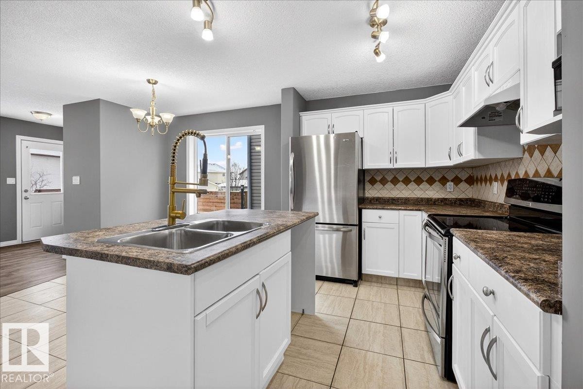 Kitchen with appliances with stainless steel finishes, dark countertops, a textured ceiling, white cabinetry, and a kitchen island with sink - 150 Acacia Circle, Leduc, AB - Indoor Photo Showing Kitchen With Double Sink