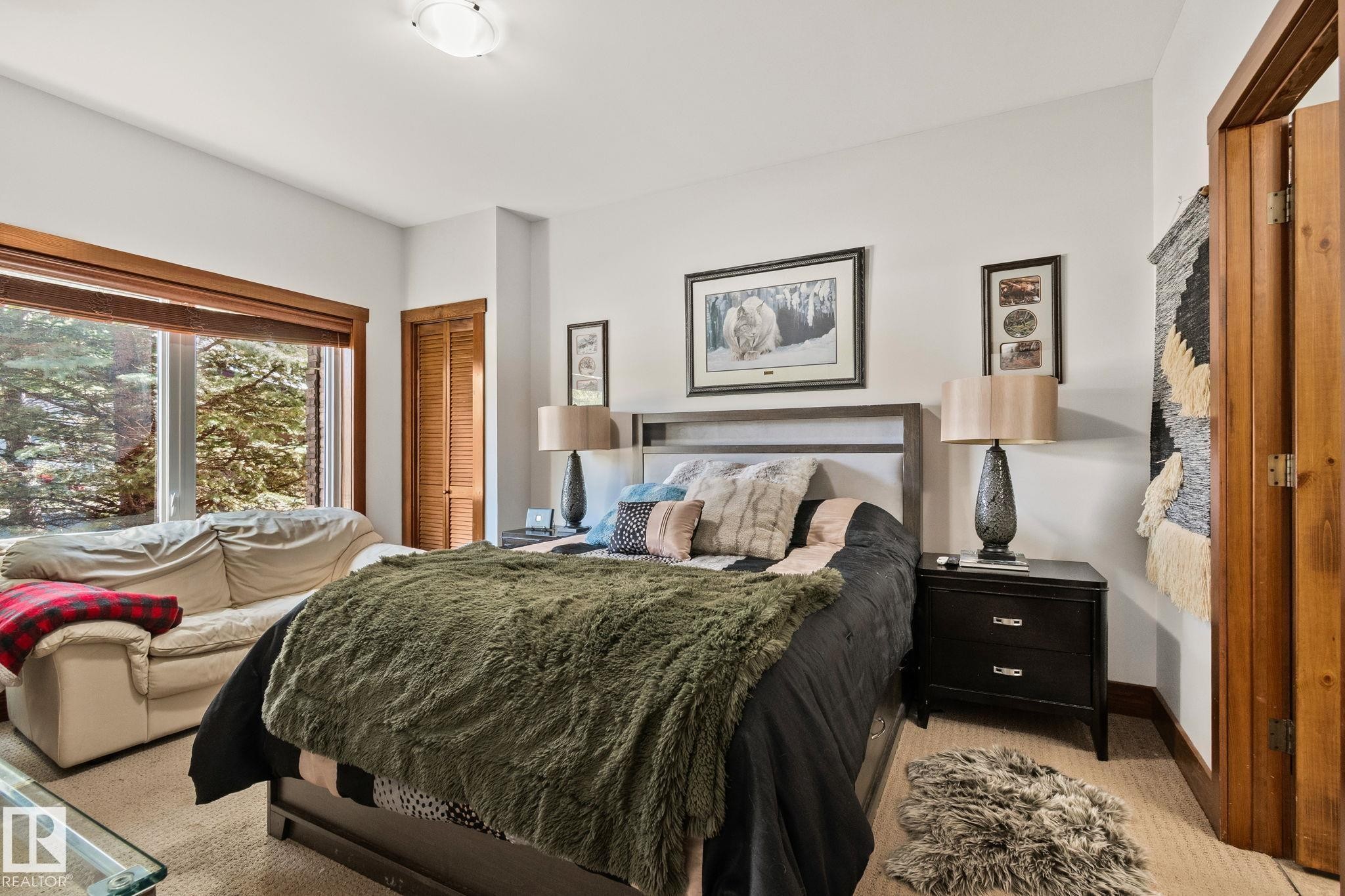Spacious room featuring a large window with wood trim, wood-finish bi-fold closet doors, wall-to-wall carpeting, and a flush-mount ceiling light fixture - 2201 Graybriar Green, Stony Plain, AB - Indoor Photo Showing Bedroom
