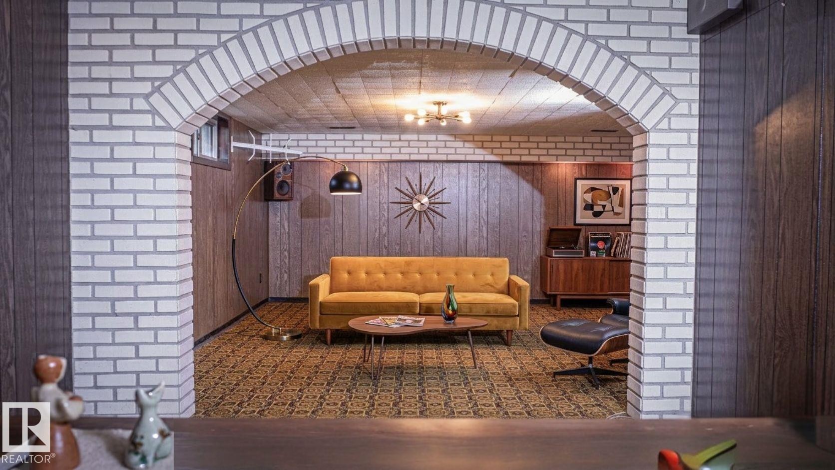 Recreational room featuring a wide arched entryway with exposed brickwork, patterned carpeting, wood-paneled walls, and a built-in ceiling light fixture - 11131 52 Street, Edmonton, AB - Indoor