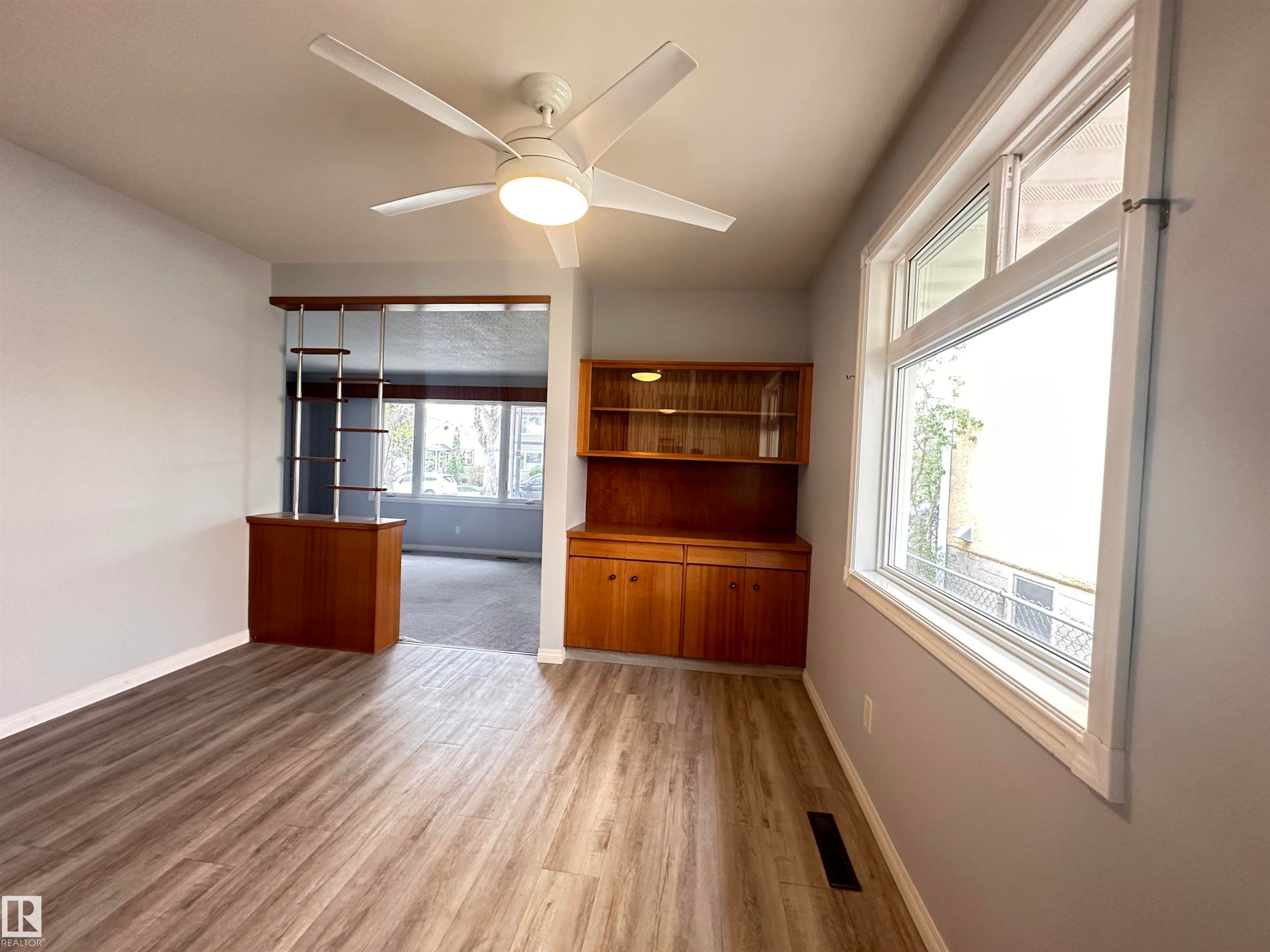 Spacious interior featuring wood-finish flooring, a contemporary ceiling fan, and a built-in wooden hutch with glass-front cabinets - 11131 52 Street, Edmonton, AB - Indoor Photo Showing Other Room