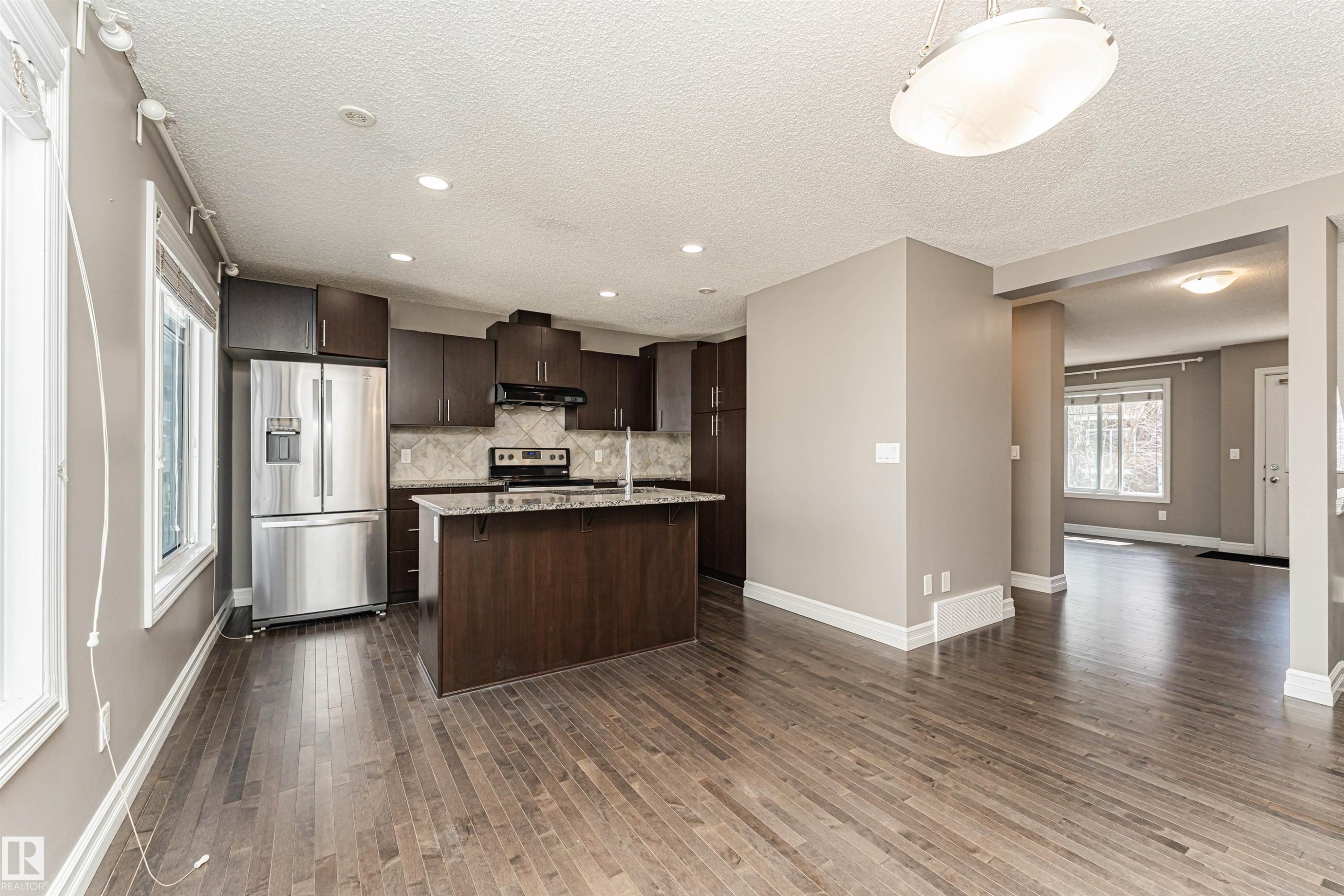 Open-concept kitchen featuring dark wood cabinetry, stainless steel appliances, a center island with a sink, and pendant lighting - #110 3625 144 Av Nw, Edmonton, AB - Indoor Photo Showing Kitchen