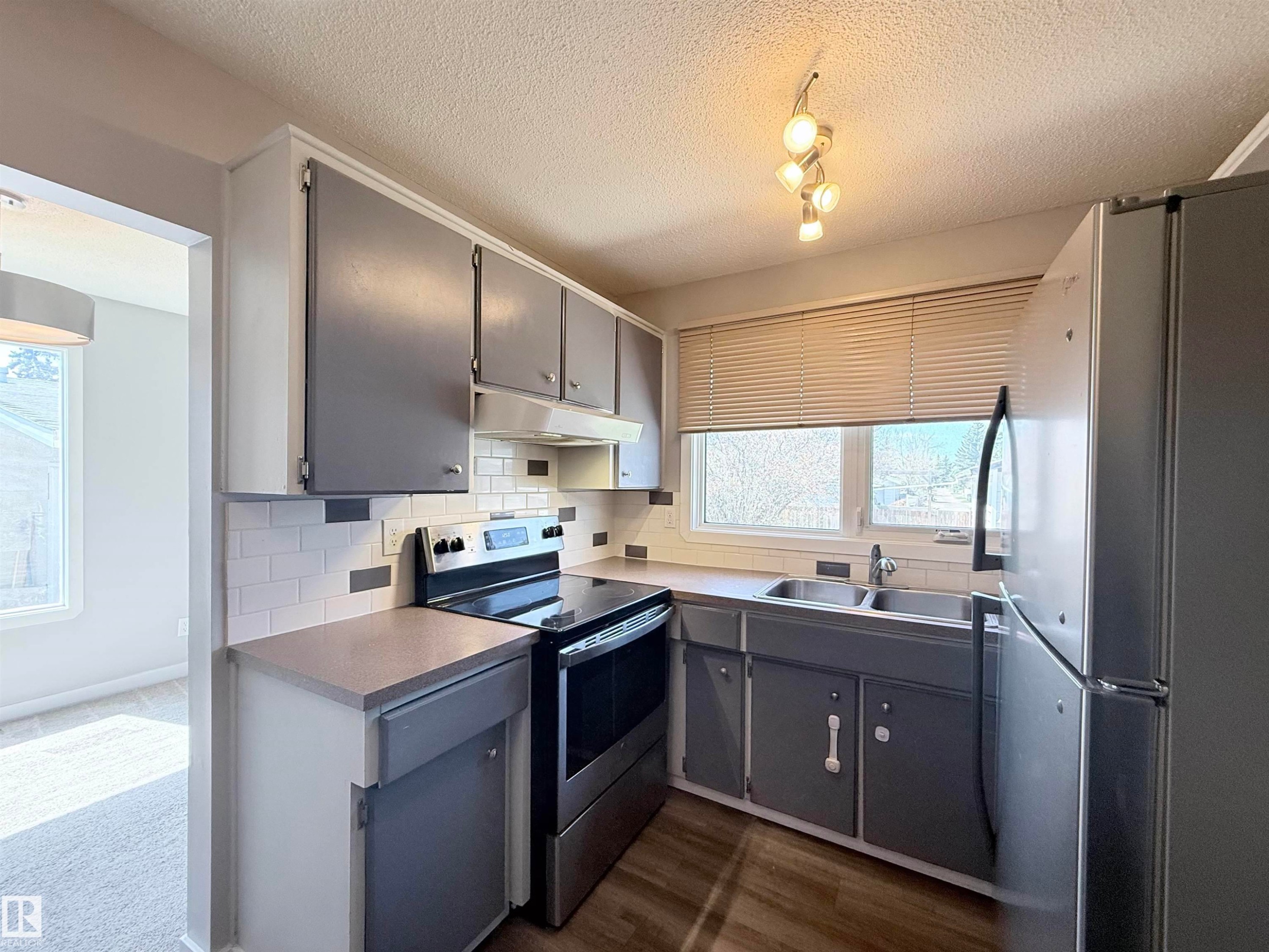 Kitchen featuring wood-finish flooring, white subway tile backsplash, stainless steel sink, track lighting, and a window with blinds - 3912 108 Street, Edmonton, AB - Indoor Photo Showing Kitchen With Double Sink