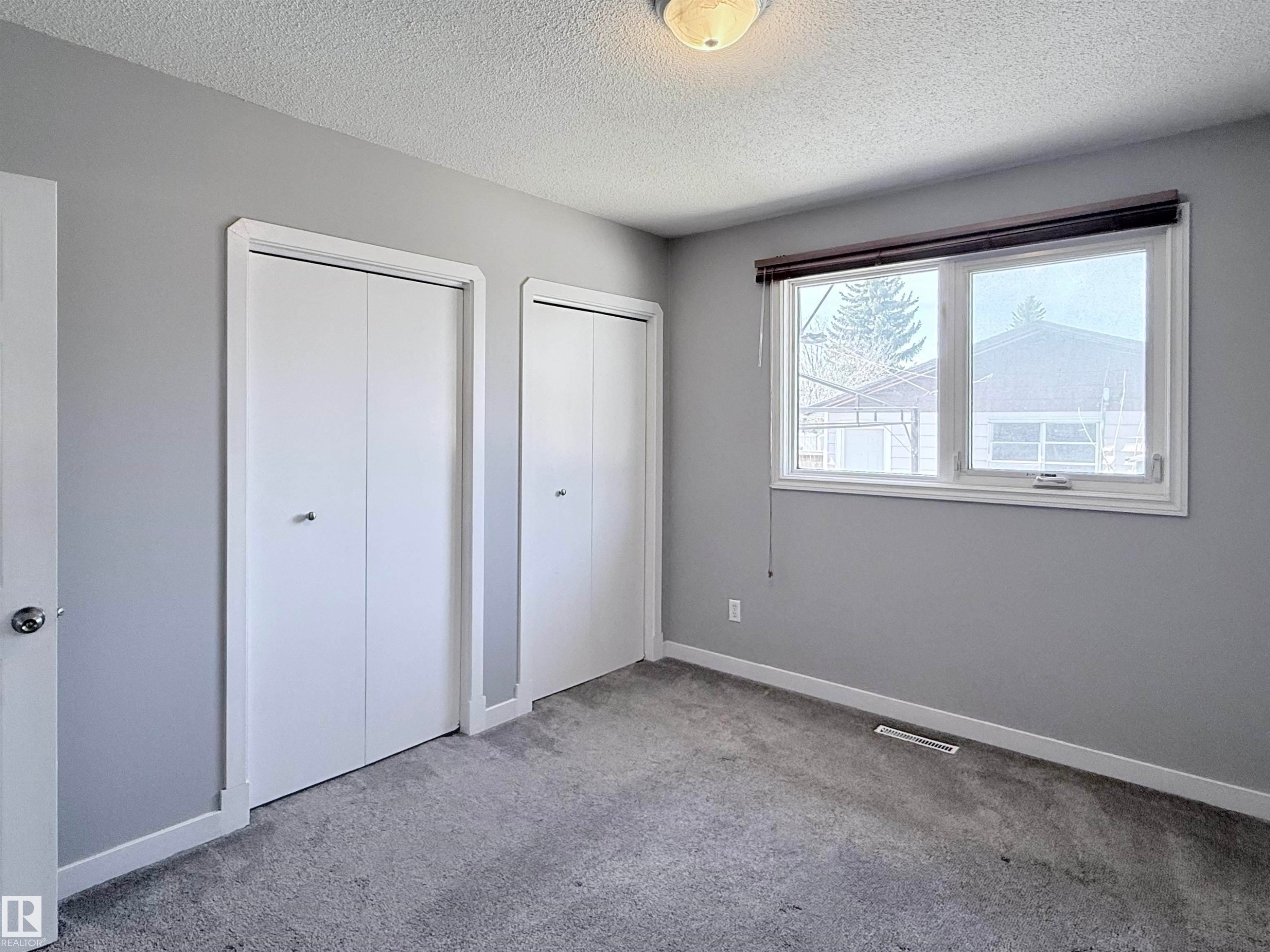 Bedroom featuring two white bi-fold closets, plush gray carpeting, and a large window with a dark roller shade - 3912 108 Street, Edmonton, AB - Indoor Photo Showing Other Room