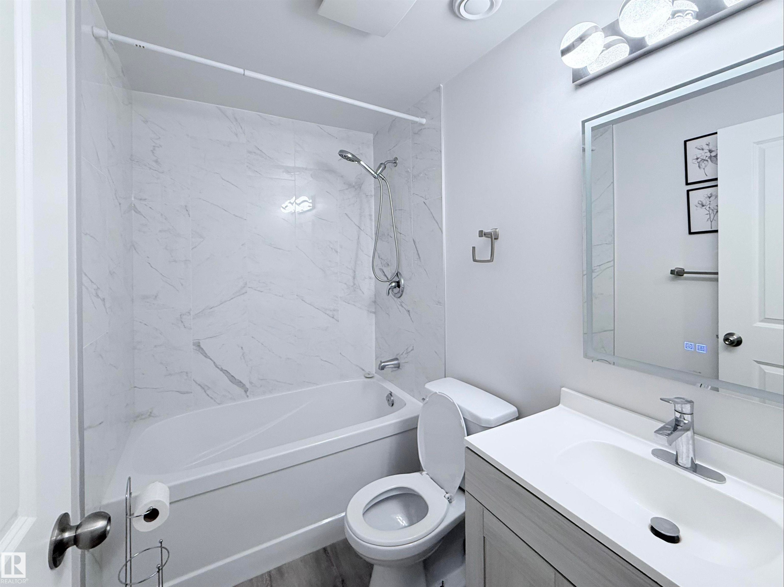 Bathroom featuring a tub and shower combination with large format marble-look tile surround, a modern vanity with integrated sink, a large backlit mirror, and wood-finish flooring - 3912 108 Street, Edmonton, AB - Indoor Photo Showing Bathroom