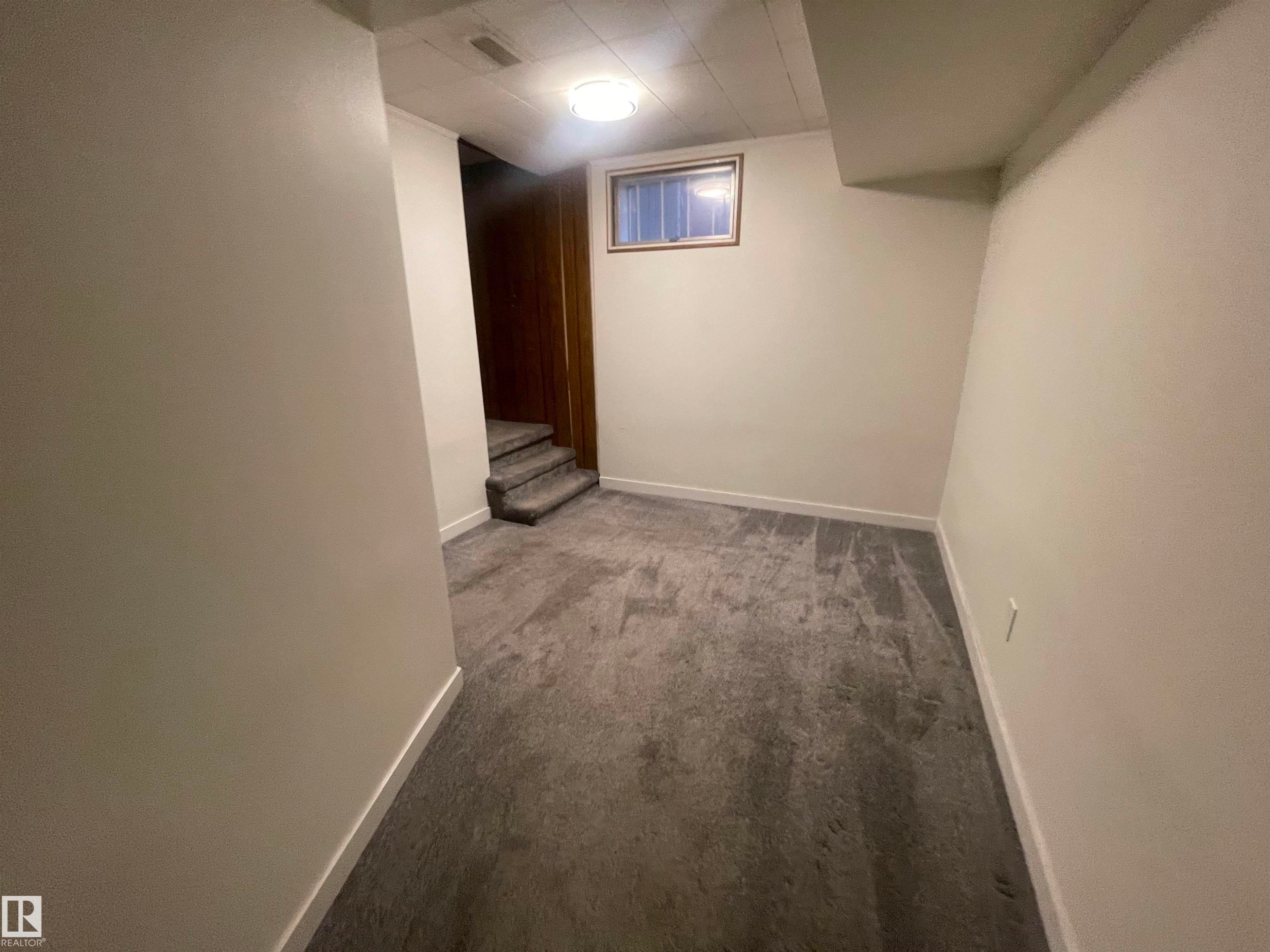 Carpeted room featuring a ceiling-mounted light fixture, a wood-trimmed window, white painted walls, white baseboards, and carpeted stairs with wood-paneled walls - 13804 132 Avenue, Edmonton, AB - Indoor Photo Showing Other Room