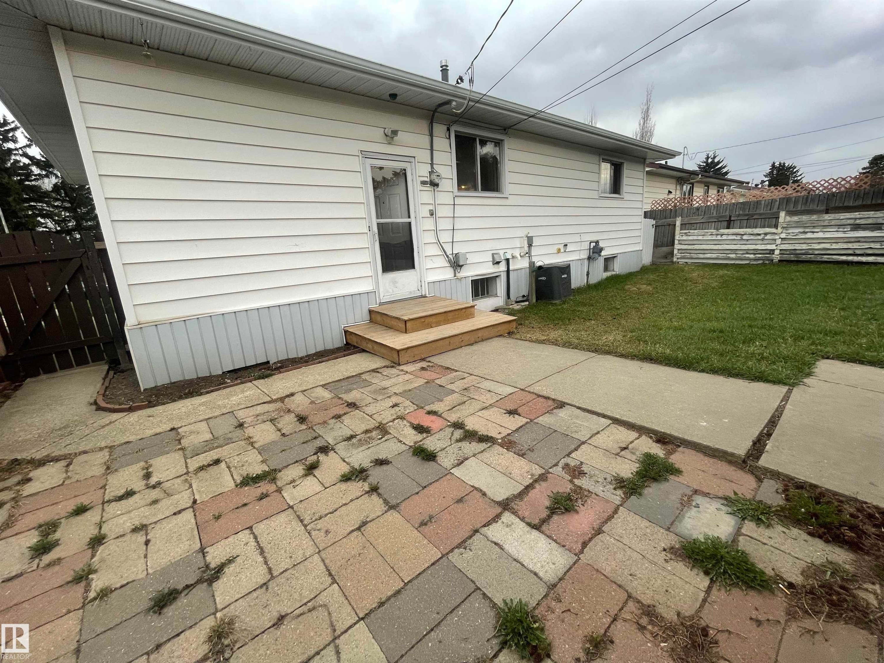 Rear exterior featuring white horizontal siding, a wooden privacy fence, a wood stair landing, and a patterned brick patio - 13804 132 Avenue, Edmonton, AB - Outdoor With Exterior