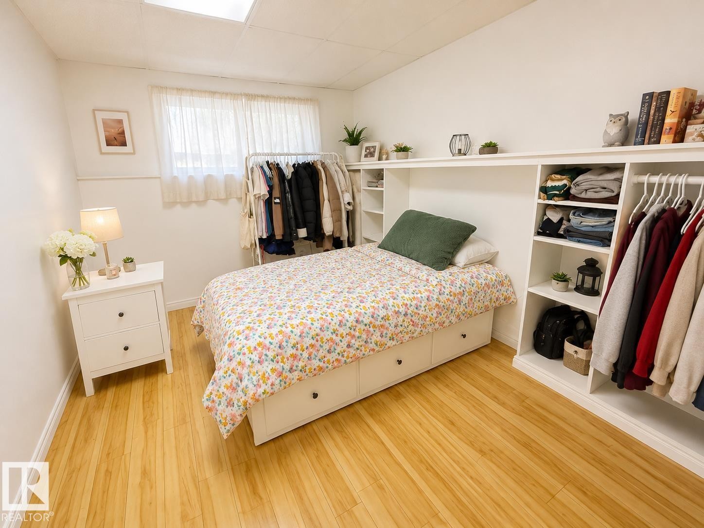 Bedroom featuring wood-finish flooring, a window with sheer curtains, a built-in storage unit with shelving and hanging space, and a bed frame with integrated drawers - 5712 101 Avenue, Edmonton, AB - Indoor Photo Showing Bedroom