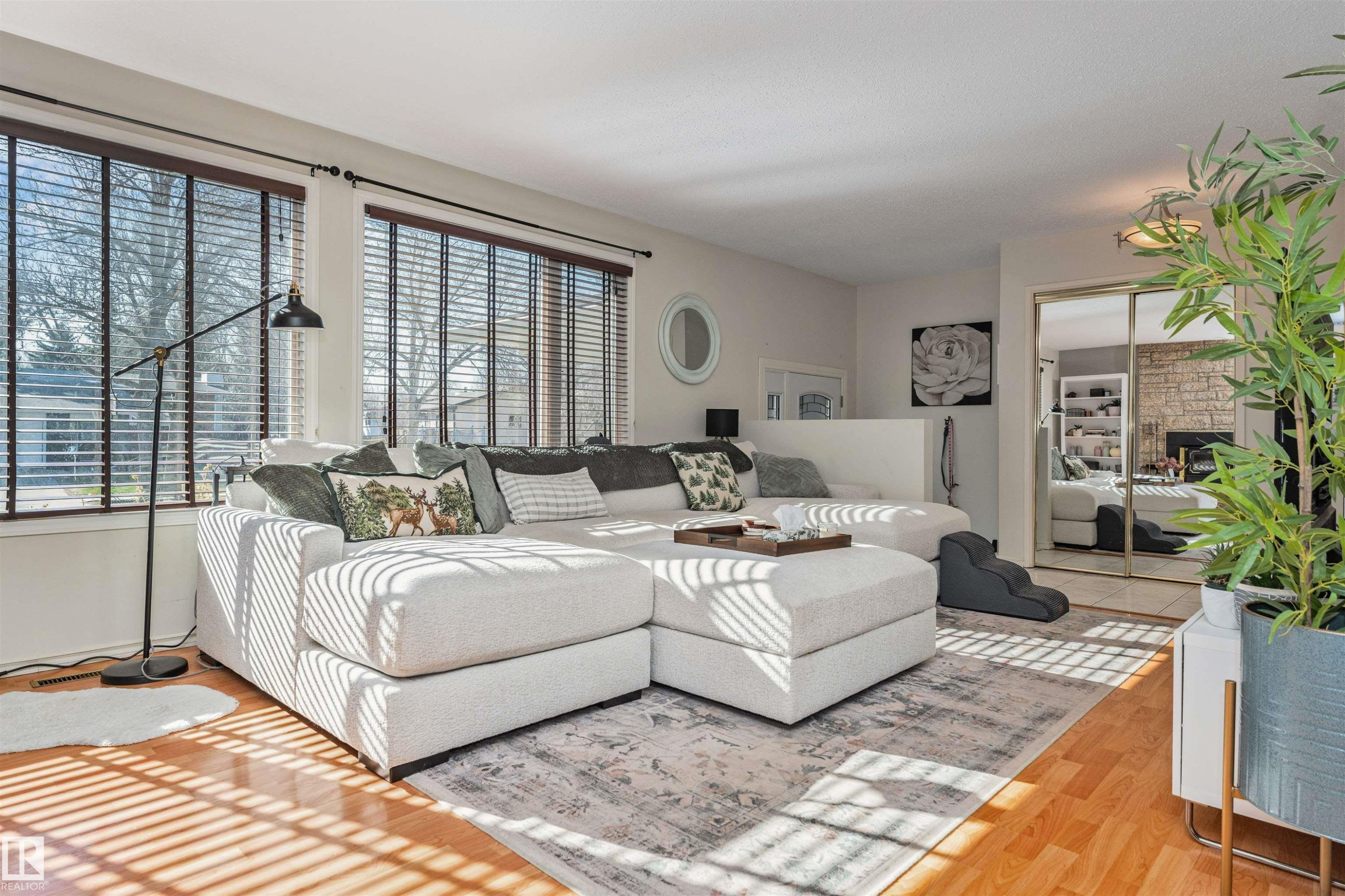 Bright living space featuring wood-finish flooring, two large windows with horizontal blinds, a large area rug, and a mirrored closet door - 49 Ash Crescent, St. Albert, AB - Indoor Photo Showing Living Room