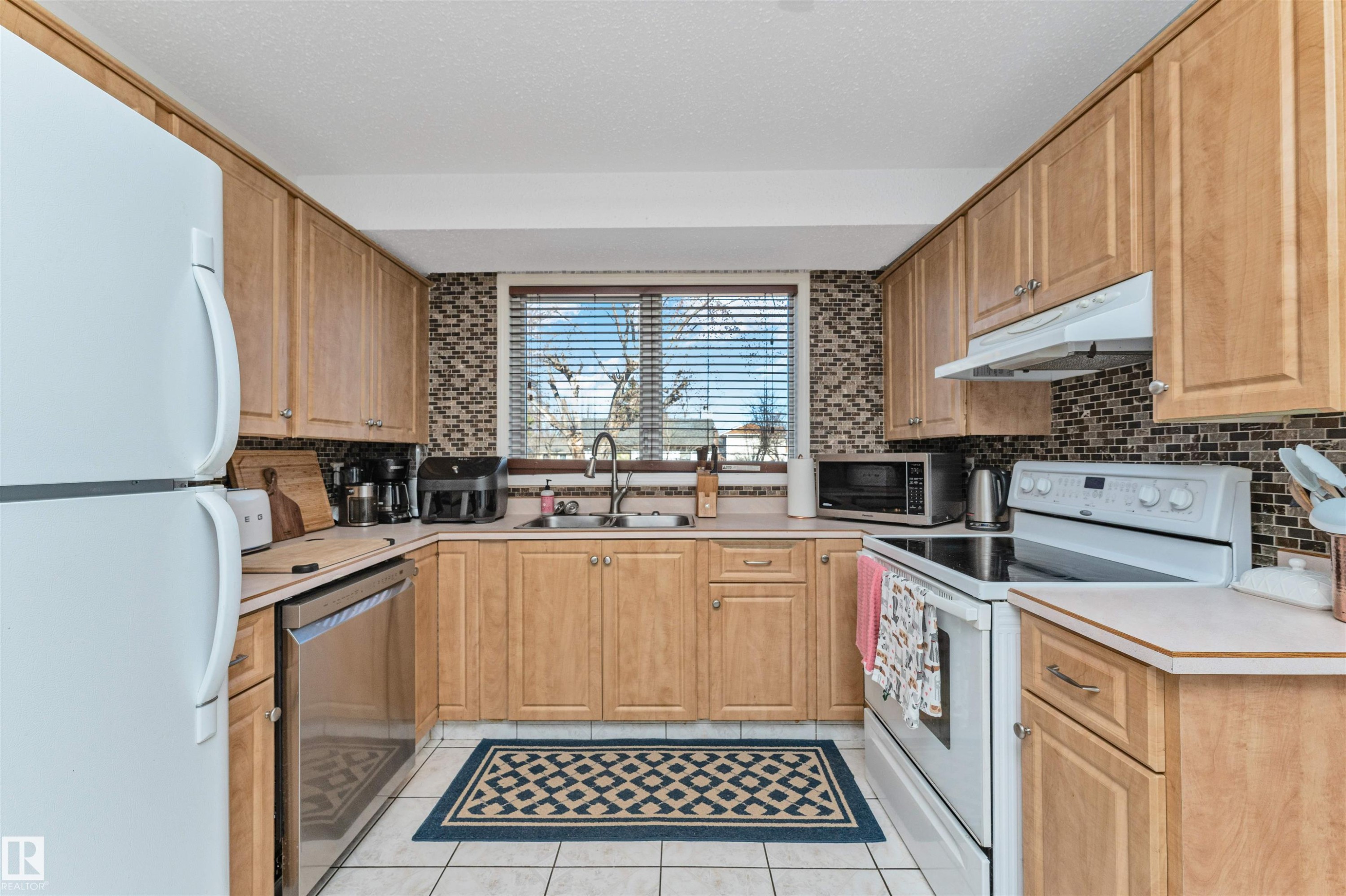 U-shaped kitchen featuring wood cabinetry, mosaic tile backsplash, white tile flooring, stainless steel dishwasher, and a window with horizontal blinds - 49 Ash Crescent, St. Albert, AB - Indoor Photo Showing Kitchen With Double Sink