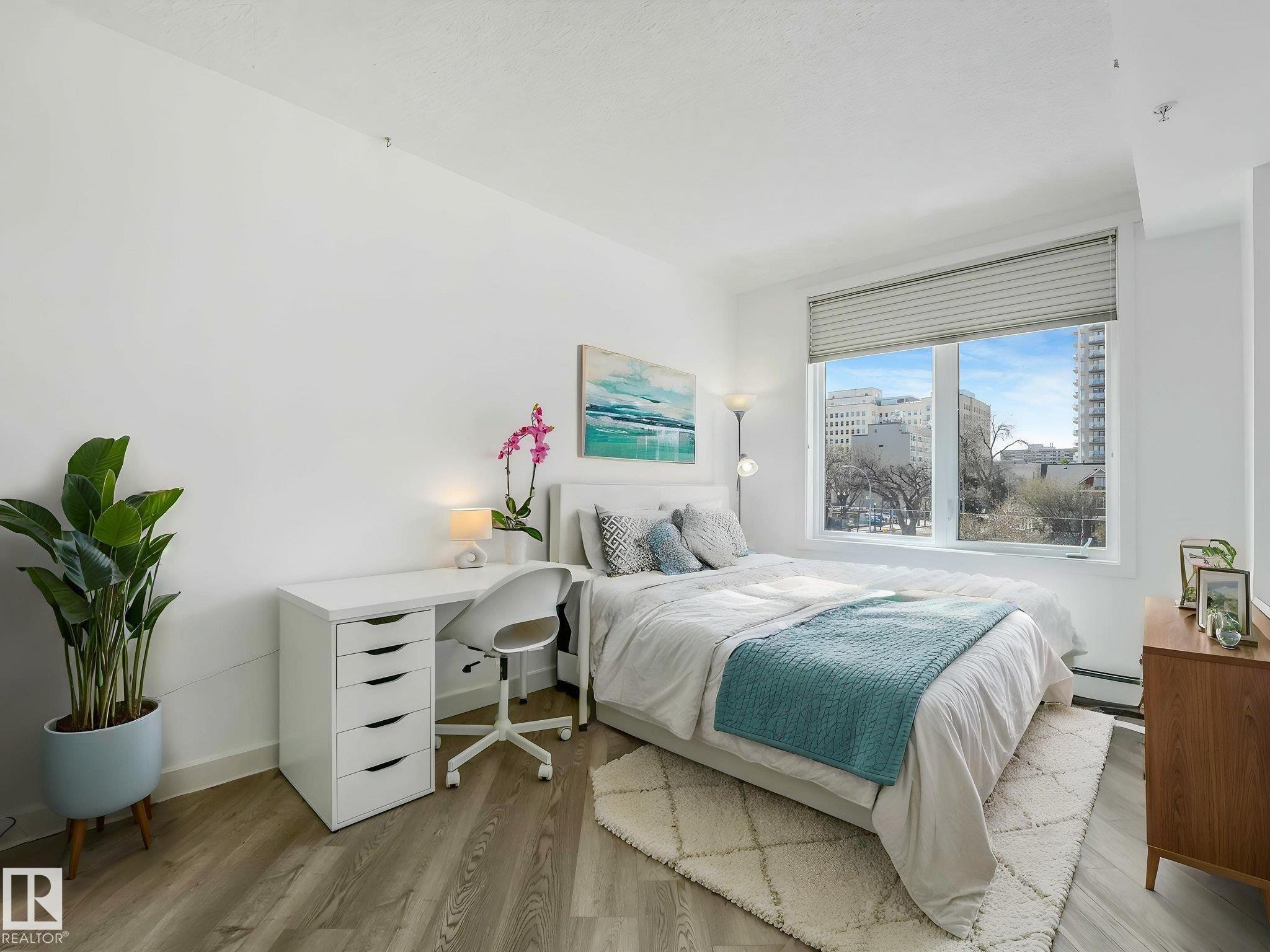 Light-filled room featuring wide window with pleat blinds, wood-finish flooring, and white painted walls - 305 10504 99 Avenue, Edmonton, AB - Indoor Photo Showing Bedroom