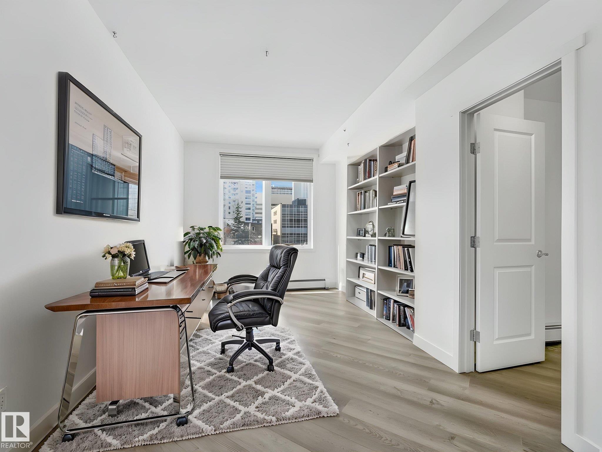 Dedicated workspace featuring light wood-finish flooring, a built-in white shelving unit, a window with roller shades, and a white two-panel door - 305 10504 99 Avenue, Edmonton, AB - Indoor
