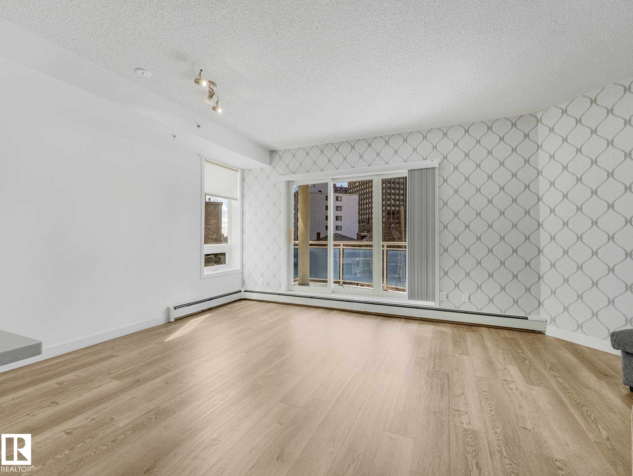 Living area featuring wood-finish flooring, a patterned accent wall, track lighting, and a balcony with cityscape scenery - 305 10504 99 Avenue, Edmonton, AB - Indoor Photo Showing Other Room