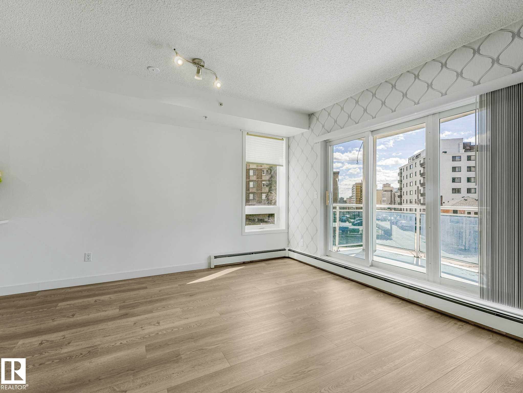 Bright living area featuring wood-finish flooring, white painted walls, a large window with a roller shade, and a three-panel glass sliding door leading to an exterior balcony - 305 10504 99 Avenue, Edmonton, AB - Indoor Photo Showing Other Room