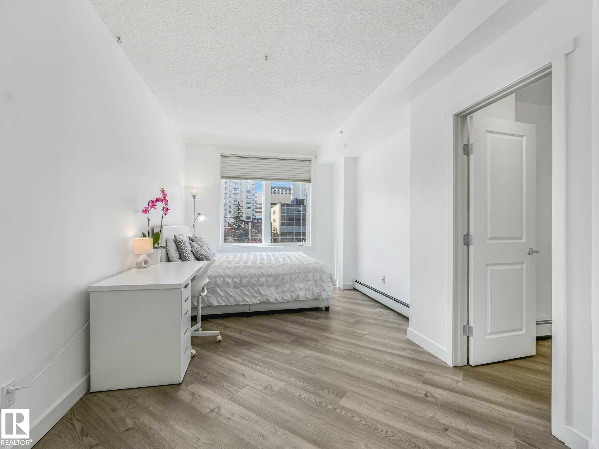 Bedroom featuring wood-finish flooring, a large window with blinds, and white baseboards - 305 10504 99 Avenue, Edmonton, AB - Indoor Photo Showing Bedroom