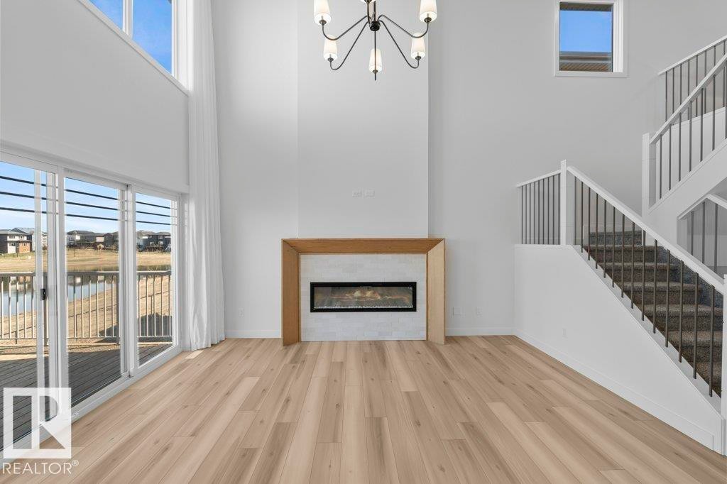 Two-story living area featuring wood-finish flooring, a contemporary fireplace with a light-toned surround and wood mantel, a multi-tiered staircase with dark balusters, a modern chandelier, and a sliding glass door opening to a deck and water feature - 19110 20A Avenue, Edmonton, AB - Indoor Photo Showing Living Room With Fireplace