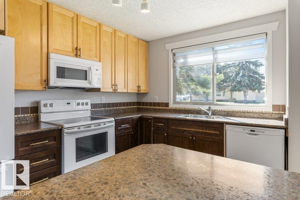 Kitchen featuring a large window, light wood upper cabinetry, dark wood lower cabinetry, white appliances, and a patterned countertop extending to a peninsula - 3736 12 Avenue, Edmonton, AB - Indoor Photo Showing Kitchen With Double Sink