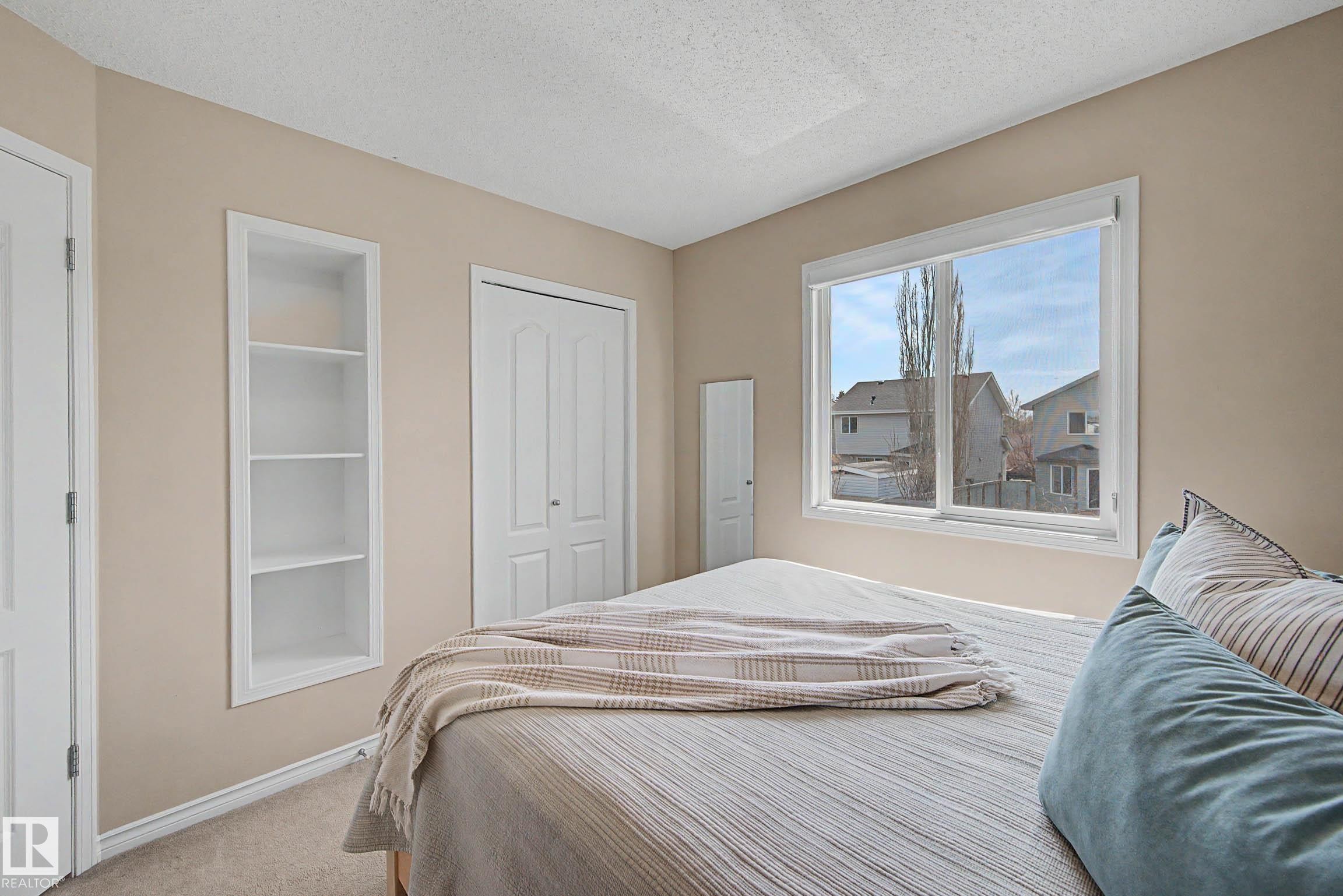 Bedroom featuring a large window, built-in shelving, two white panel doors, and neutral-toned carpeting - 7723 7A Avenue, Edmonton, AB - Indoor Photo Showing Bedroom