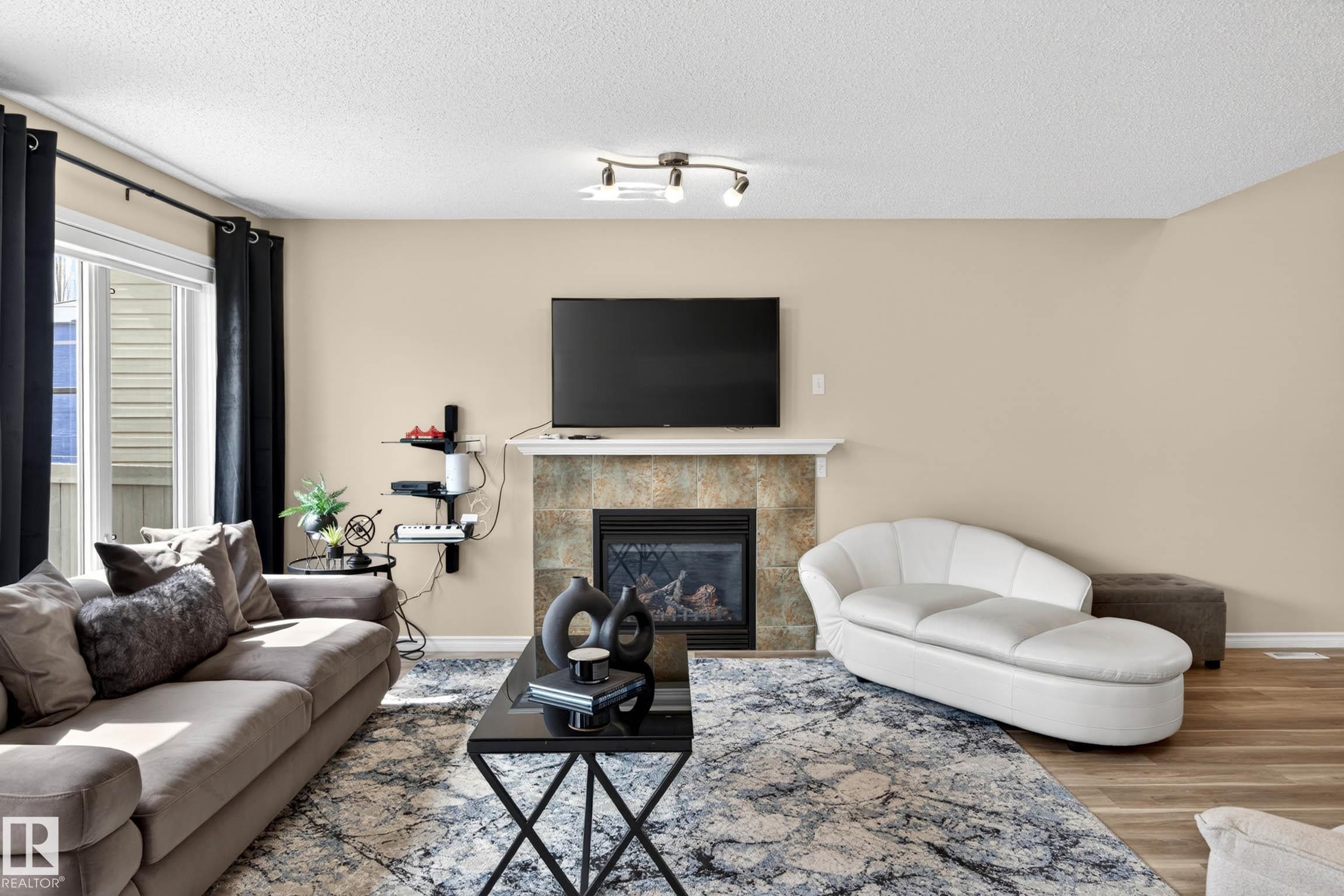 Living area featuring a tiled fireplace with mantel, built-in electric insert, and a large window with vertical blinds - 7723 7A Avenue, Edmonton, AB - Indoor Photo Showing Living Room With Fireplace