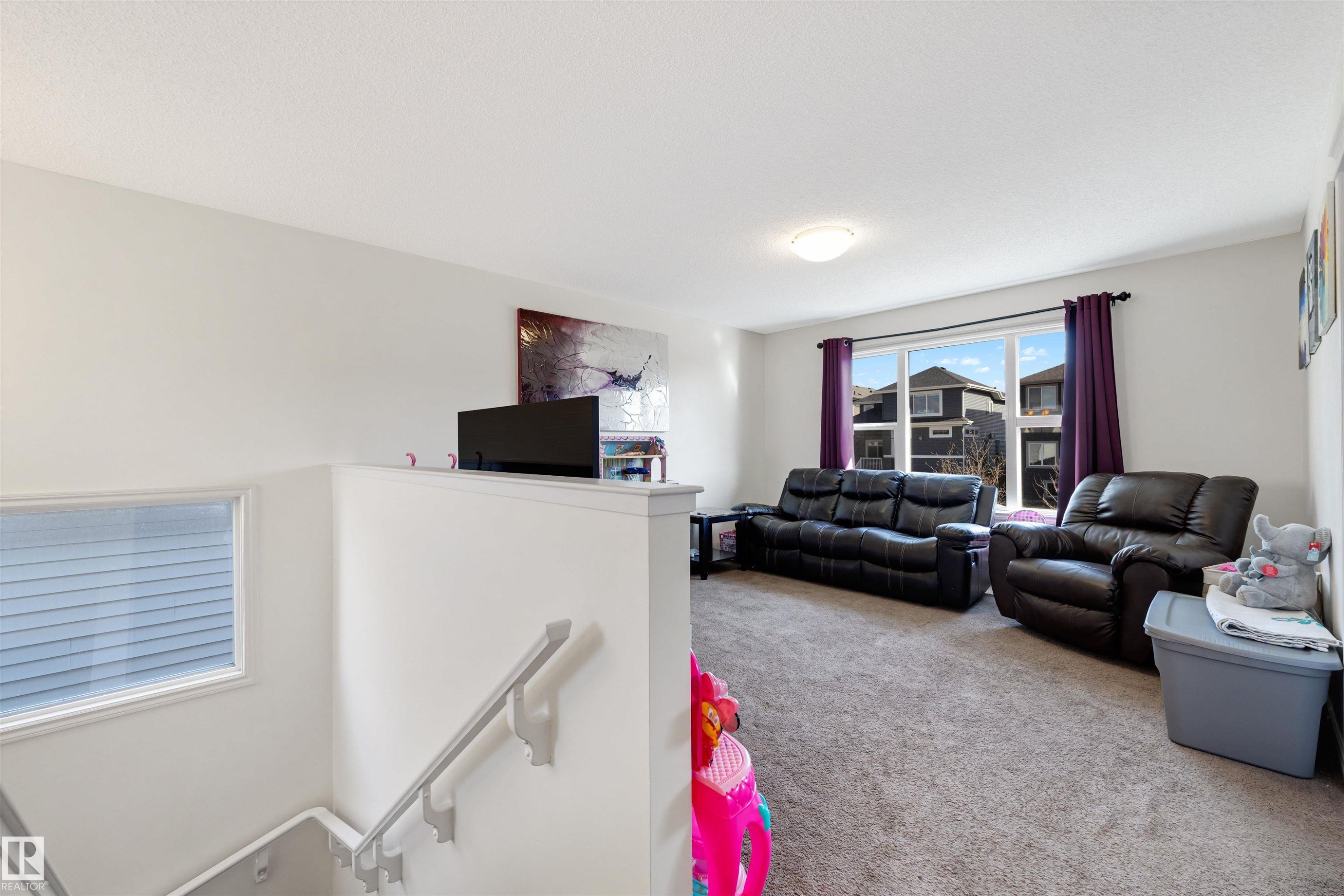 Carpeted living area featuring a large window with purple drapes, white walls, and a flush mount ceiling light - 6115 18 Avenue, Edmonton, AB - Indoor