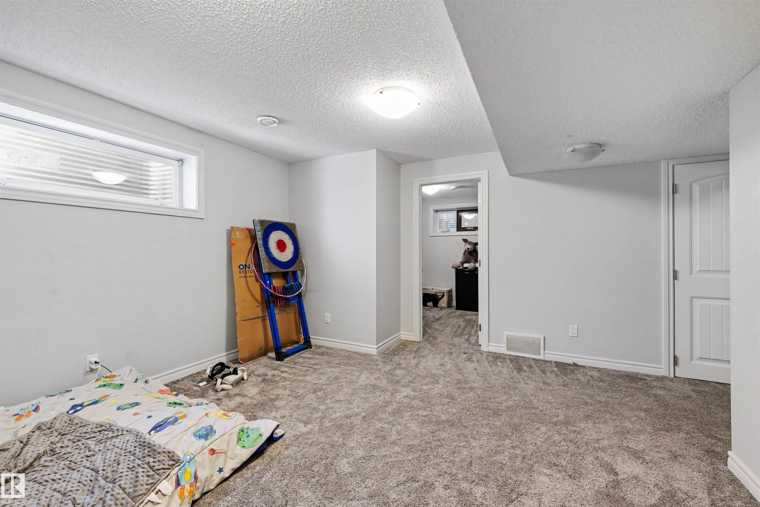 Spacious carpeted room featuring a white trim window with blinds, ceiling-mounted light fixtures, a paneled interior door, and light gray wall paint - 6115 18 Avenue, Edmonton, AB - Indoor Photo Showing Bedroom