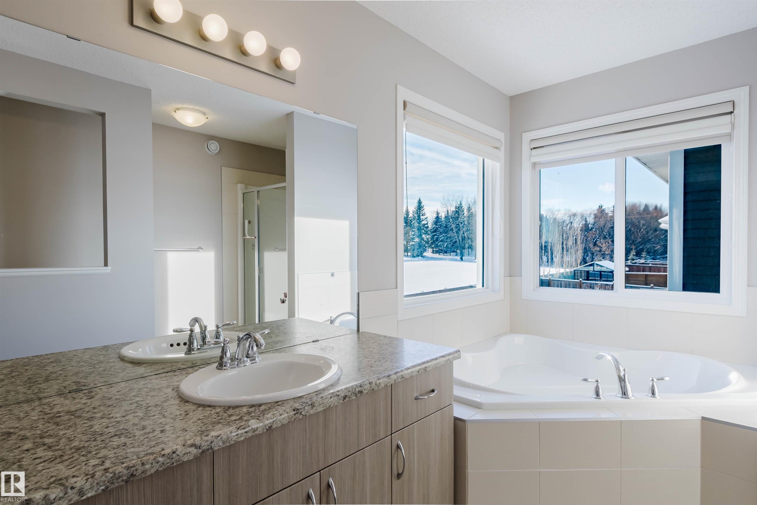 Spacious bathroom featuring a built-in corner bathtub, dual windows, a double vanity with stone-finish countertop, and integrated sinks with chrome faucets - 16627 18 Avenue, Edmonton, AB - Indoor Photo Showing Bathroom