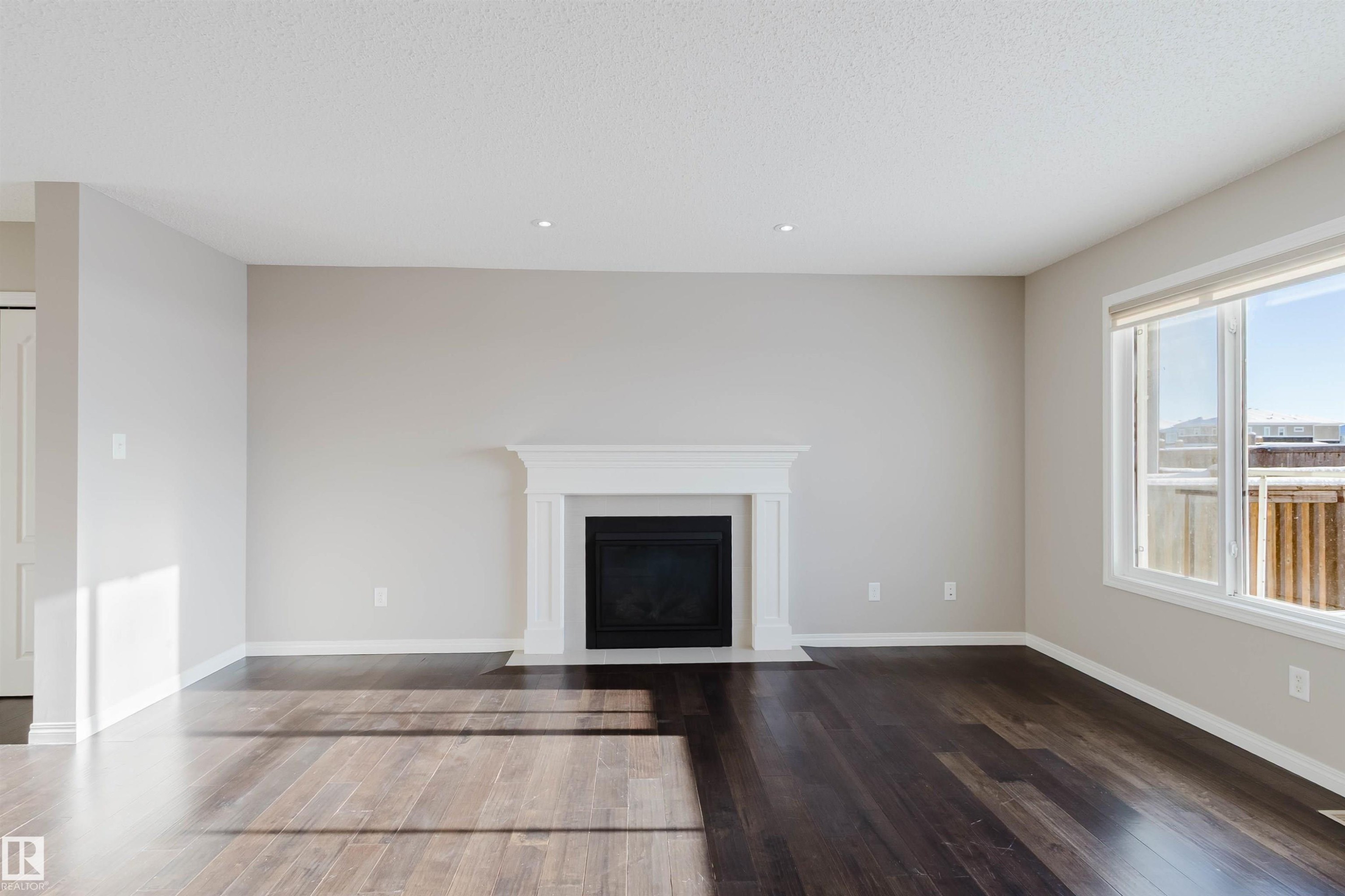 Spacious room featuring dark wood-finish flooring, a white mantel fireplace, large windows with white trim, recessed lighting, and light neutral wall colors - 16627 18 Avenue, Edmonton, AB - Indoor Photo Showing Living Room With Fireplace