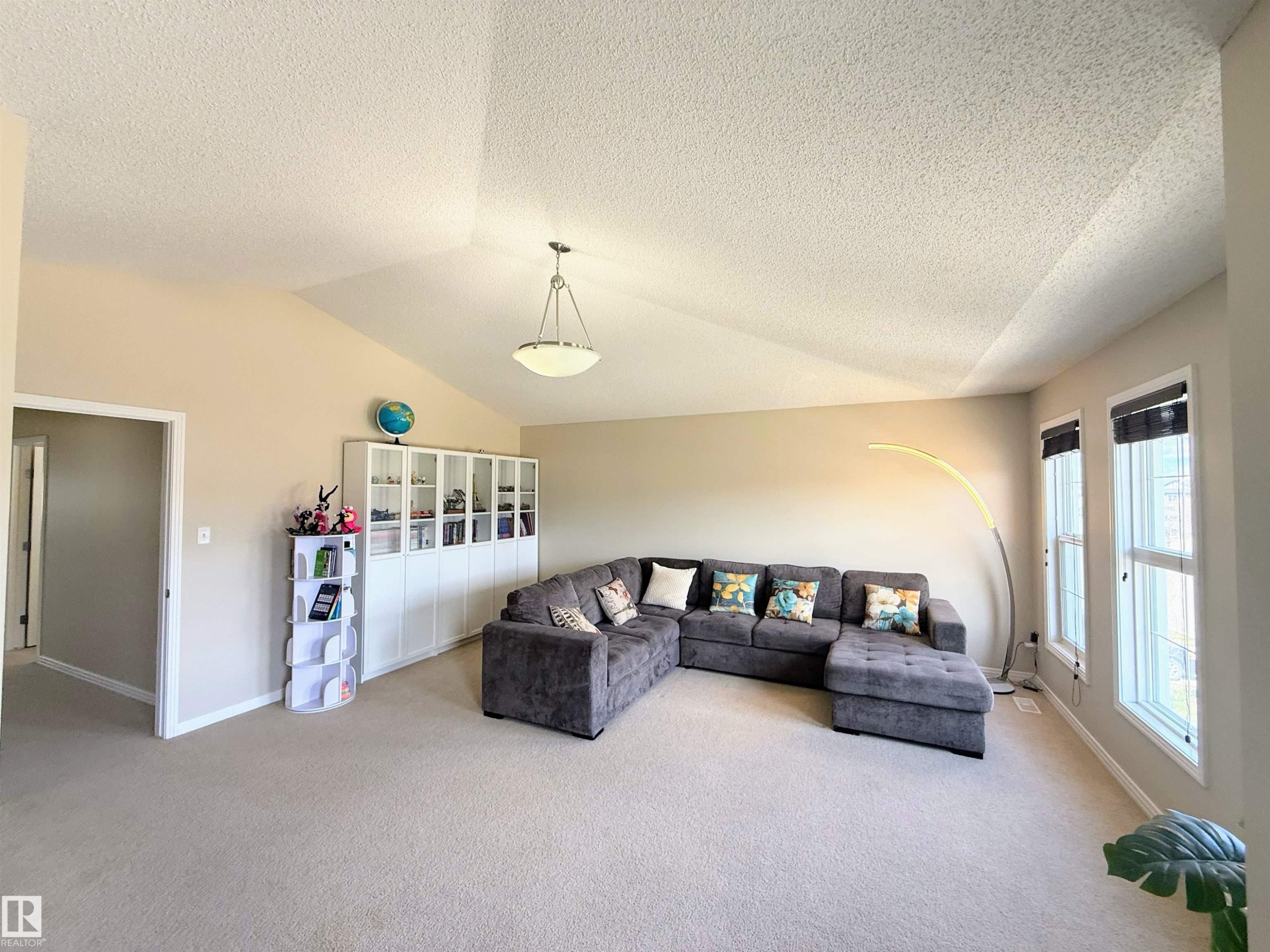 Spacious living area featuring vaulted ceilings, light-toned carpeting, a built-in wall unit with glass-fronted cabinets, and multiple windows with roller shades - 3538 Claxton Crescent, Edmonton, AB - Indoor