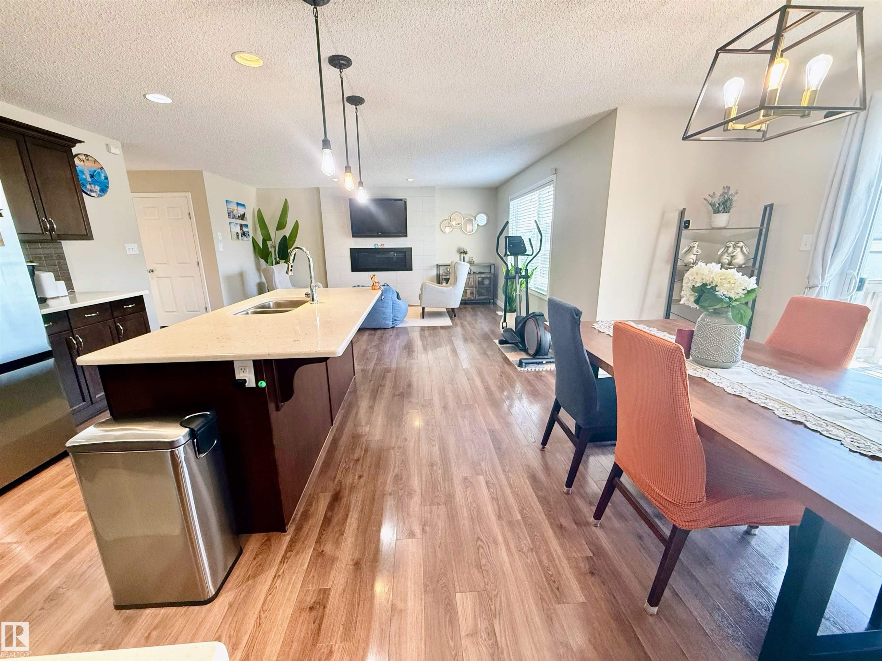 Open-concept living space featuring wood-finish flooring, a kitchen island with integrated sink, pendant lighting, dark cabinetry, and a built-in fireplace - 3538 Claxton Crescent, Edmonton, AB - Indoor Photo Showing Other Room