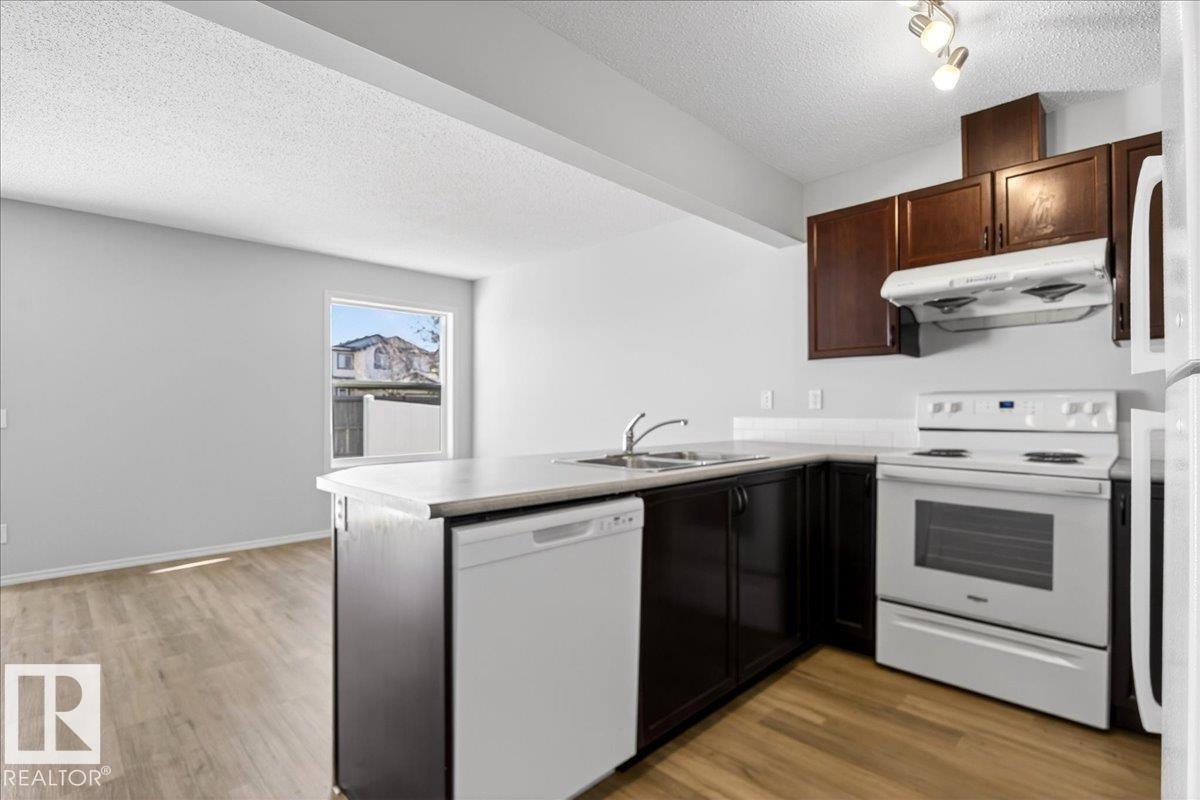 Kitchen featuring wood-finish flooring, dark wood cabinetry, light-toned countertops, white appliances, and a double basin sink - #7 18004 89 St Nw, Edmonton, AB - Indoor Photo Showing Kitchen With Double Sink