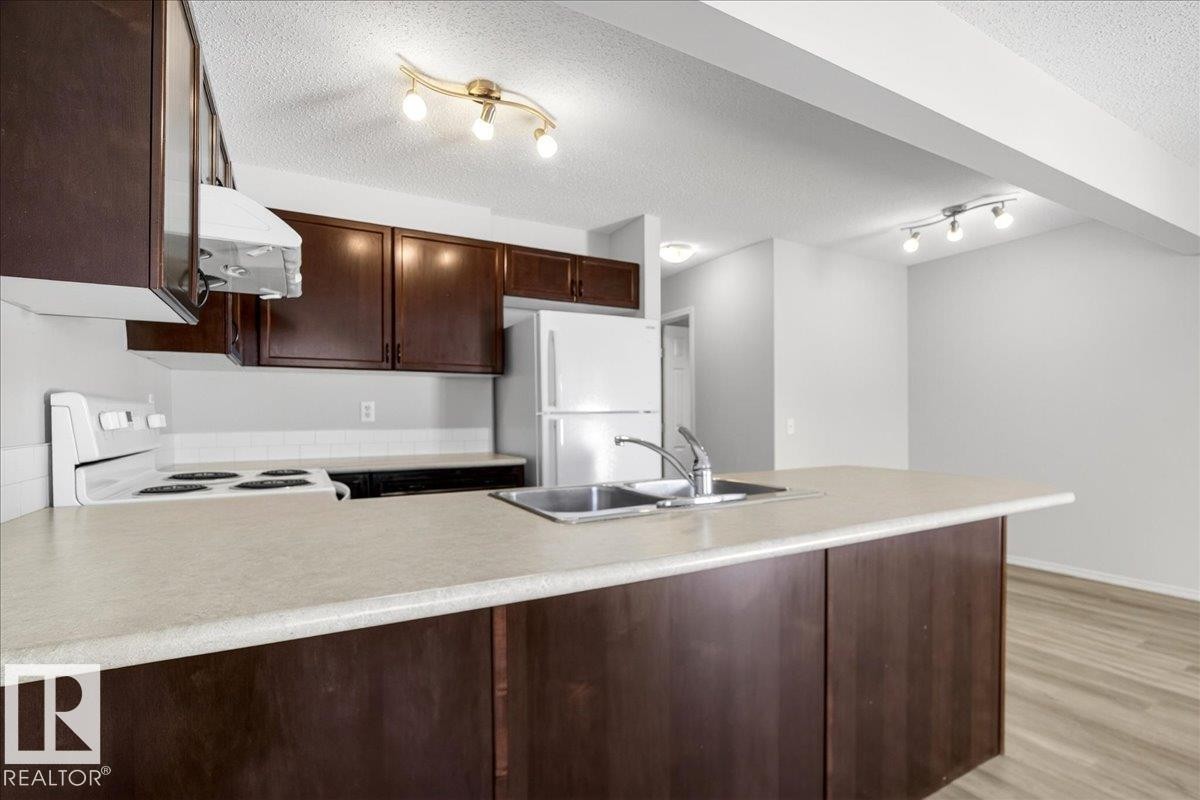 Kitchen featuring a peninsula with integrated sink, dark wood-finish cabinetry, light-toned countertops, and a white tile backsplash - #7 18004 89 St Nw, Edmonton, AB - Indoor Photo Showing Kitchen