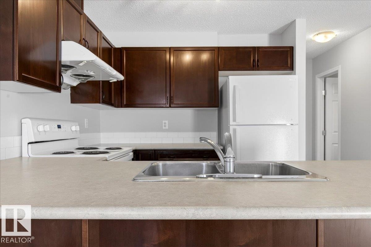Kitchen featuring rich wood-finish cabinetry, light-toned countertops, a stainless steel double-basin sink with a chrome faucet, a white electric range, and a white refrigerator - #7 18004 89 St Nw, Edmonton, AB - Indoor Photo Showing Kitchen With Double Sink