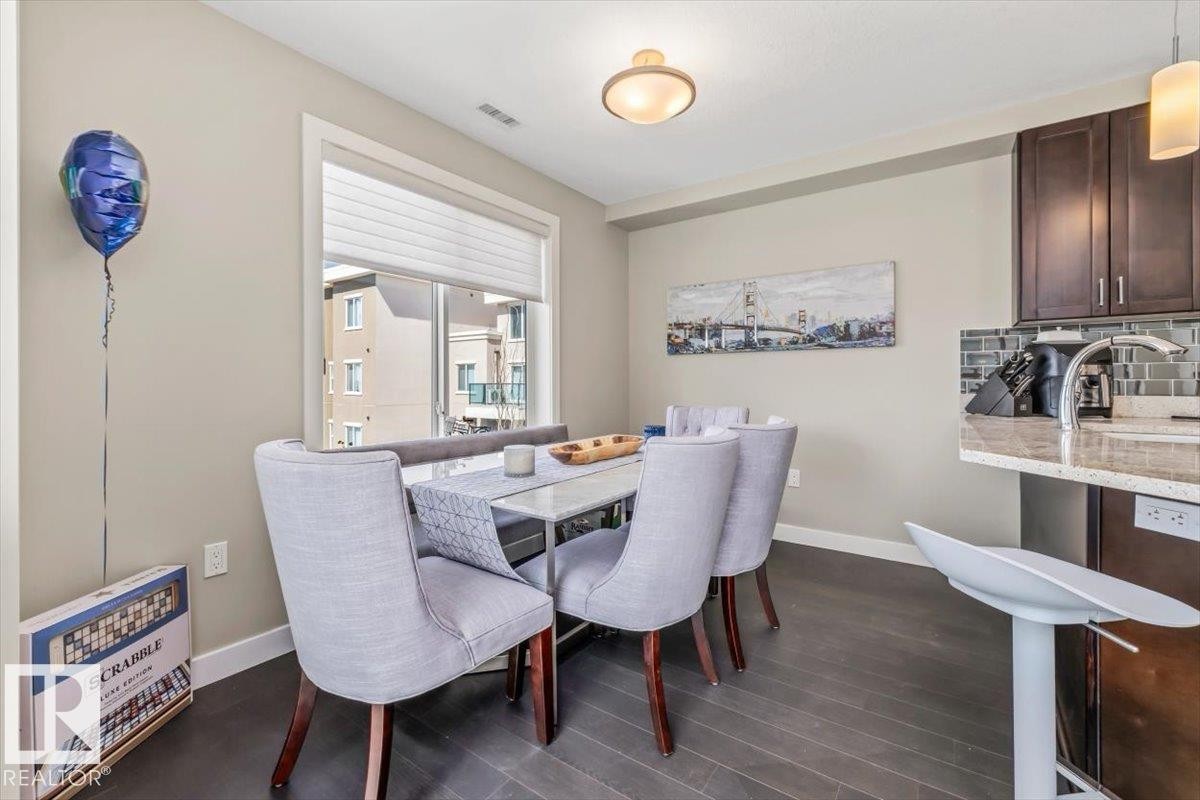 Modern kitchen featuring a granite-topped island with breakfast bar seating, dark wood-finish cabinetry, stainless steel appliances, and a subway tile backsplash - 513 1230 Windermere Way, Edmonton, AB